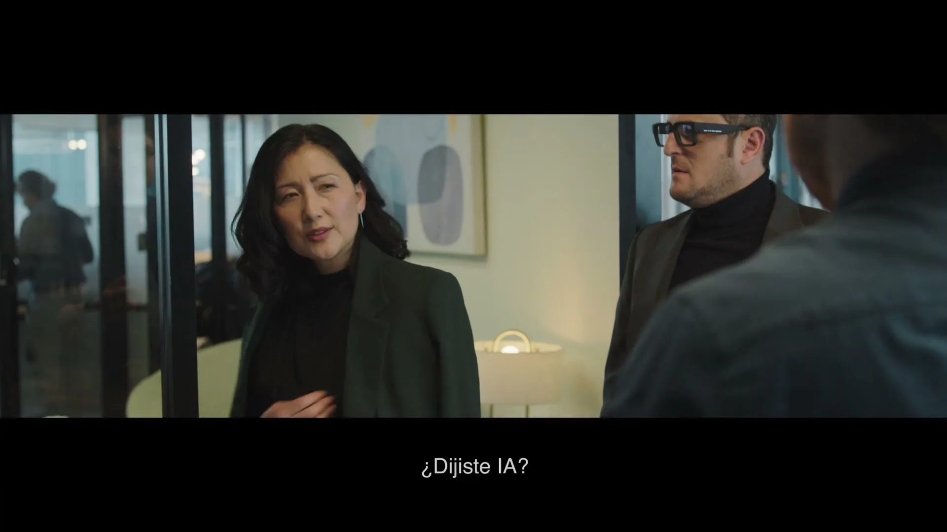 A woman in a dark green blazer turns to face the camera, looking surprised, while a man in glasses stands behind her in an office setting.