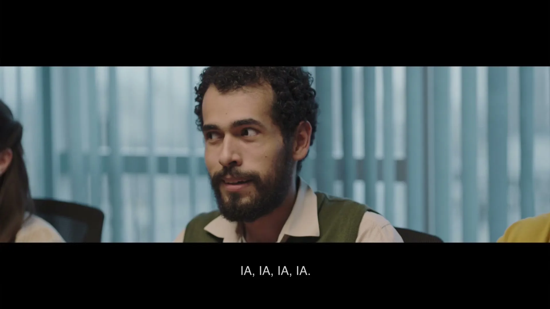 A man with a beard and curly hair, wearing a green vest over a collared shirt, looks around a conference room with a curious expression.