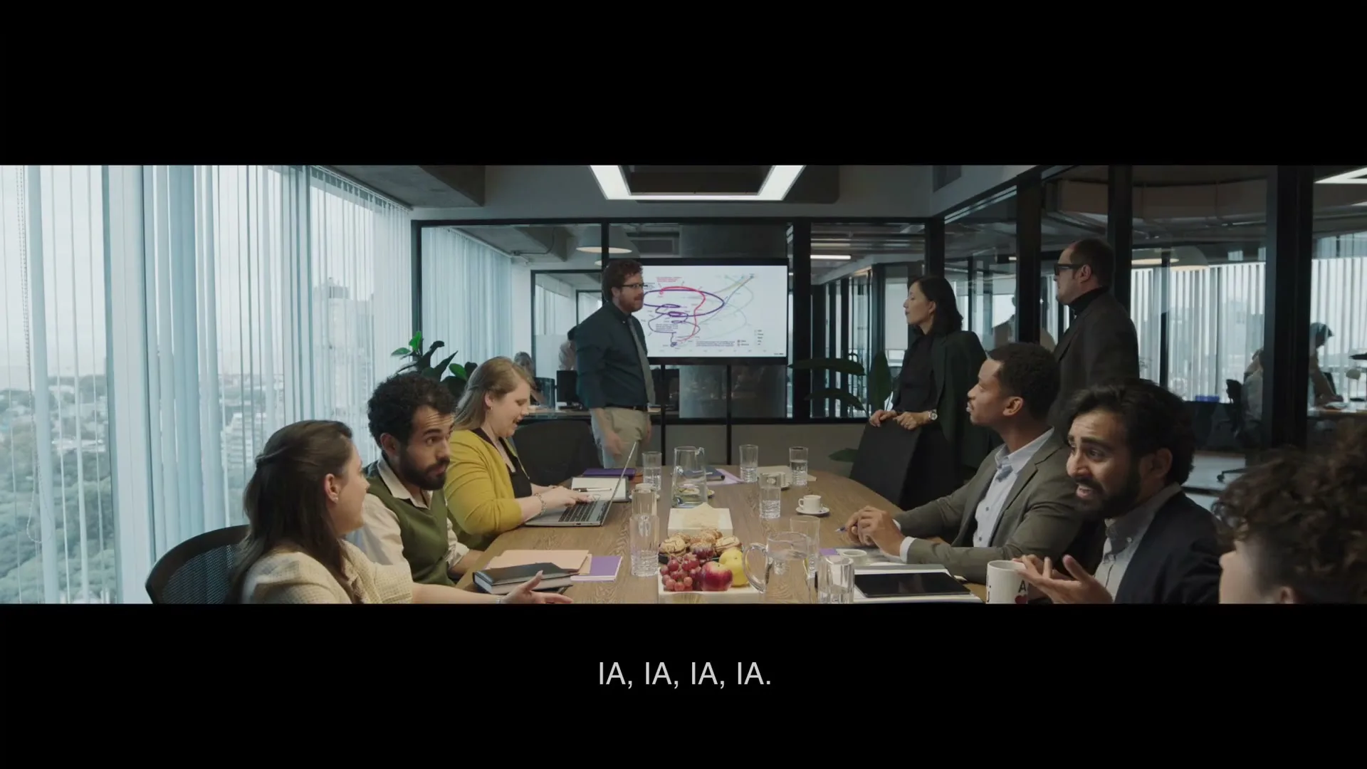 A diverse group of business people sit around a conference table in a modern office, listening to a man presenting on a screen.