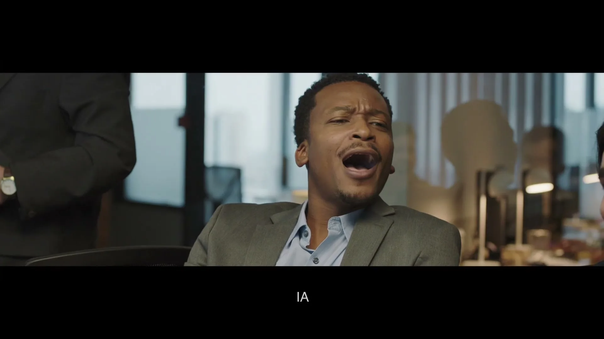 A Black man in a suit reacts with a pained expression, then smiles, sitting in an office with colleagues nearby.