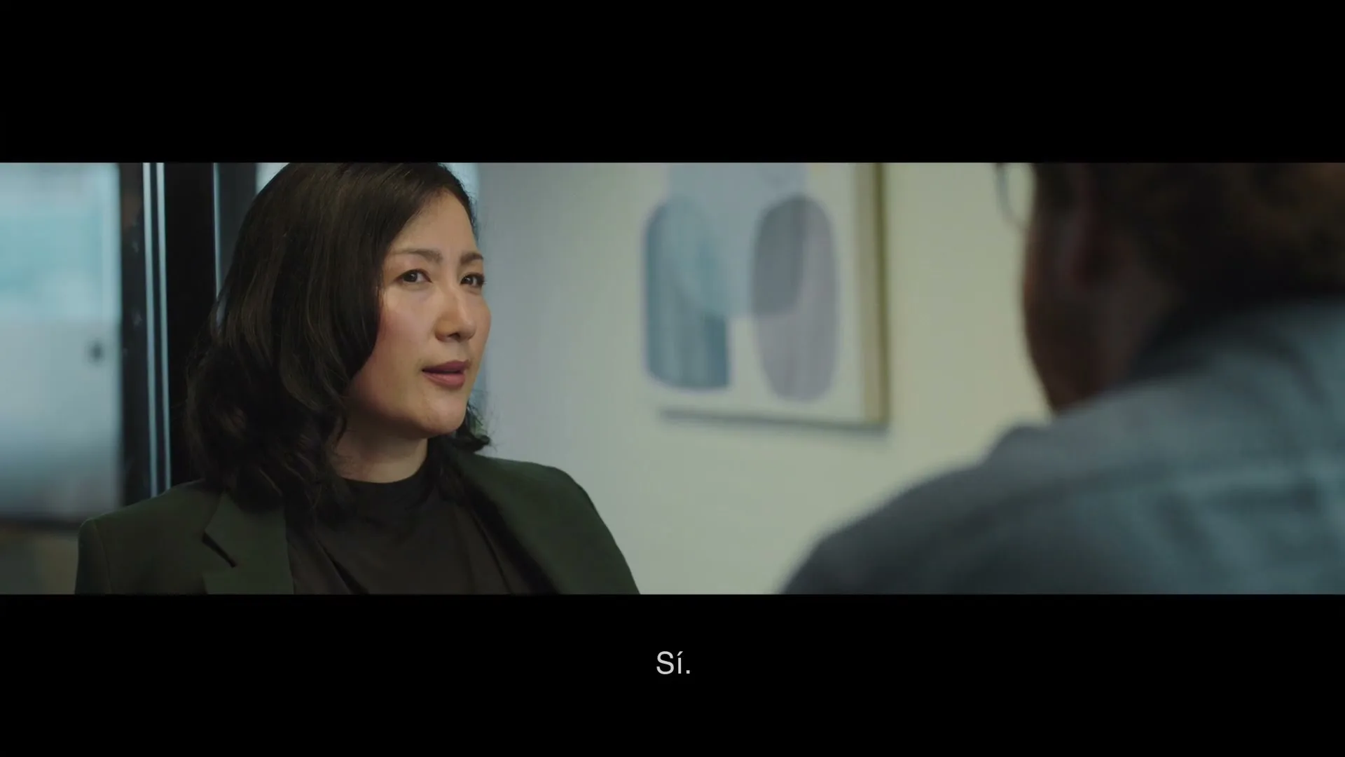 A woman with dark hair in a dark green blazer talks to an unseen person in an office setting.