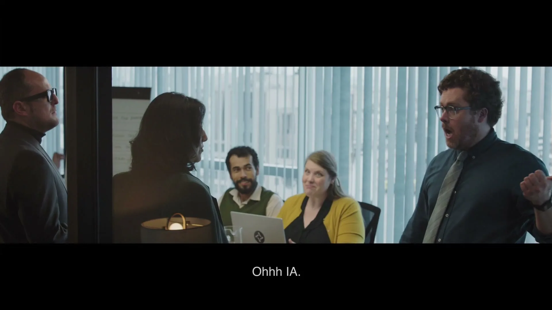 A group of office workers in a meeting room, with one man on the right gesturing excitedly while others look on, some smiling.