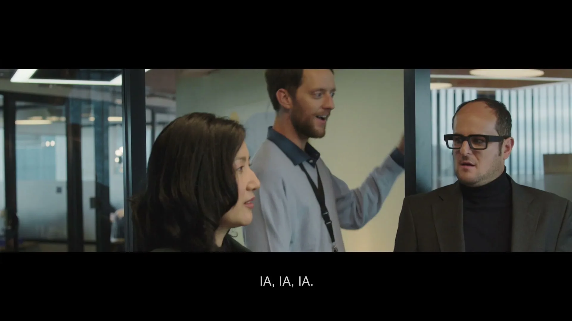 Three colleagues, two men and one woman, stand in an office, looking at a screen and talking, with the man in the middle gesturing.