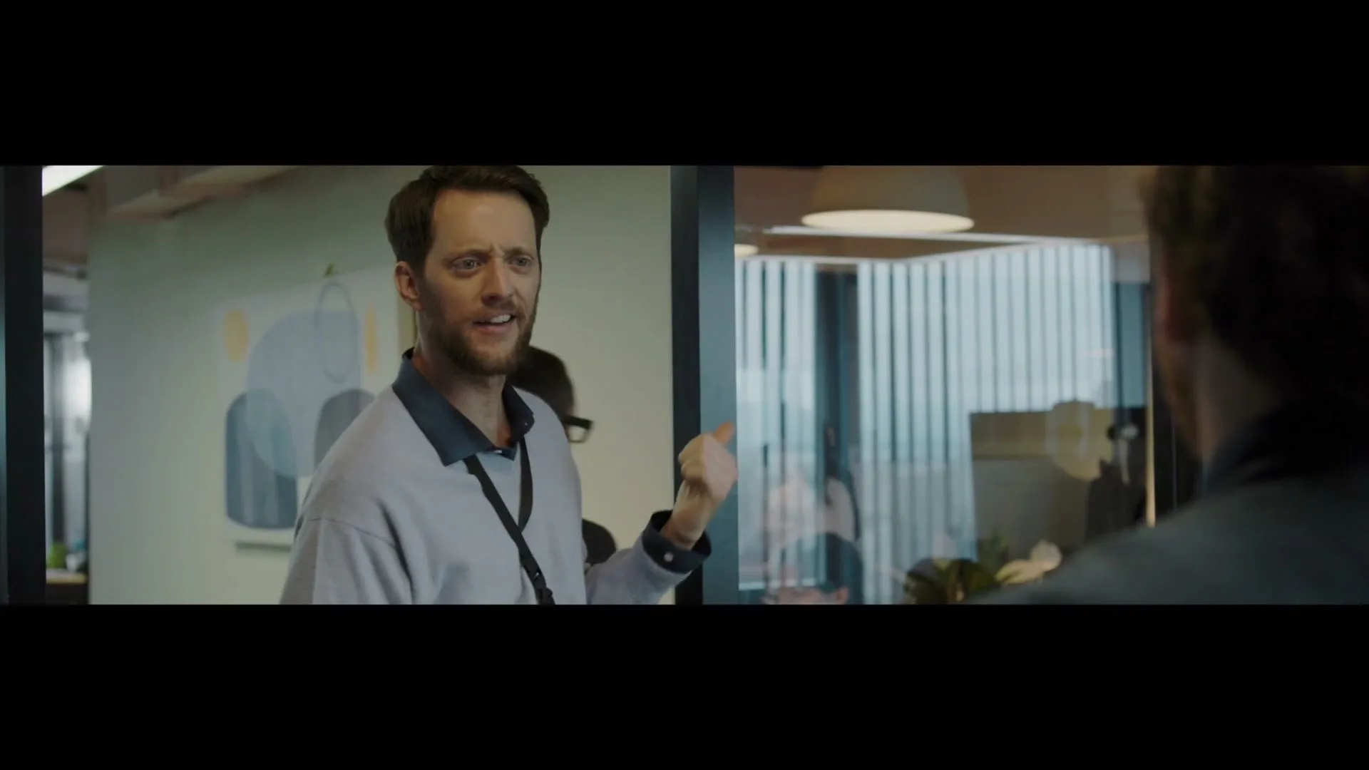 A man in a light blue sweatshirt and lanyard talks to someone off-camera, gesturing with his hand in a modern office setting.