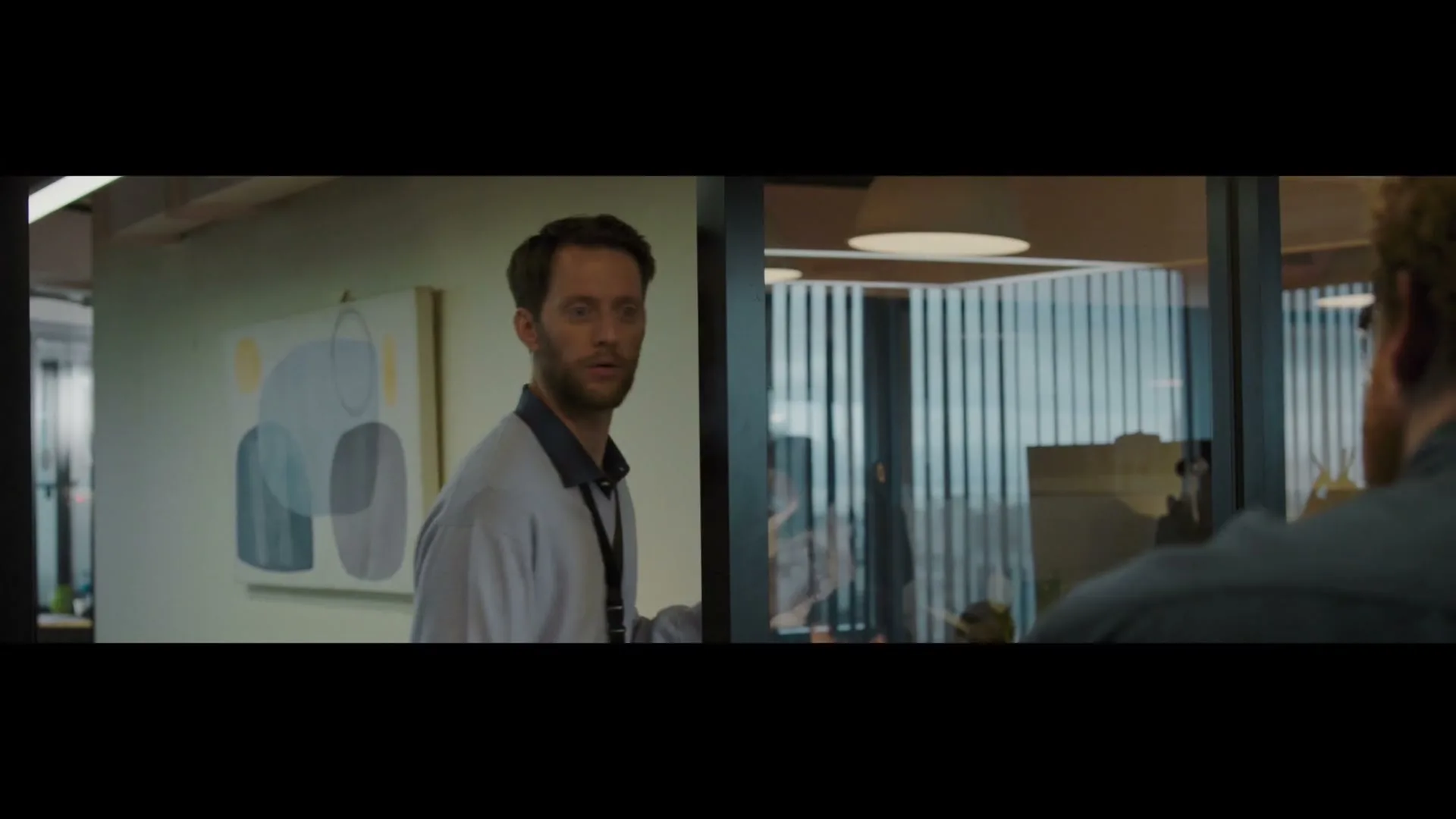 A man with a beard and light blue polo shirt looks surprised as he watches someone walk past him in an office.