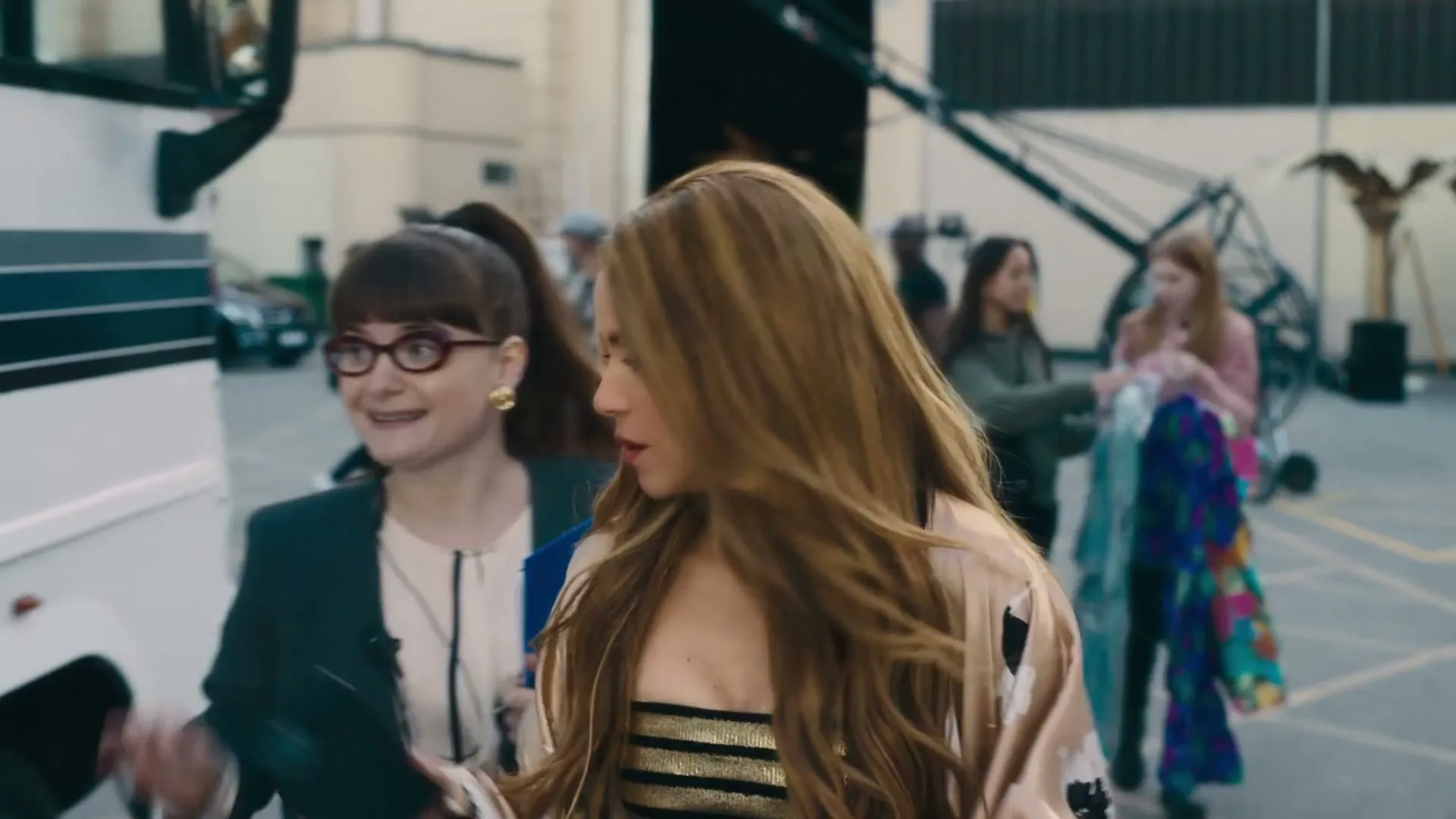 Shakira, a woman with long blonde hair and a gold top, walks past a white bus with a female assistant in glasses and a suit, on a film set.