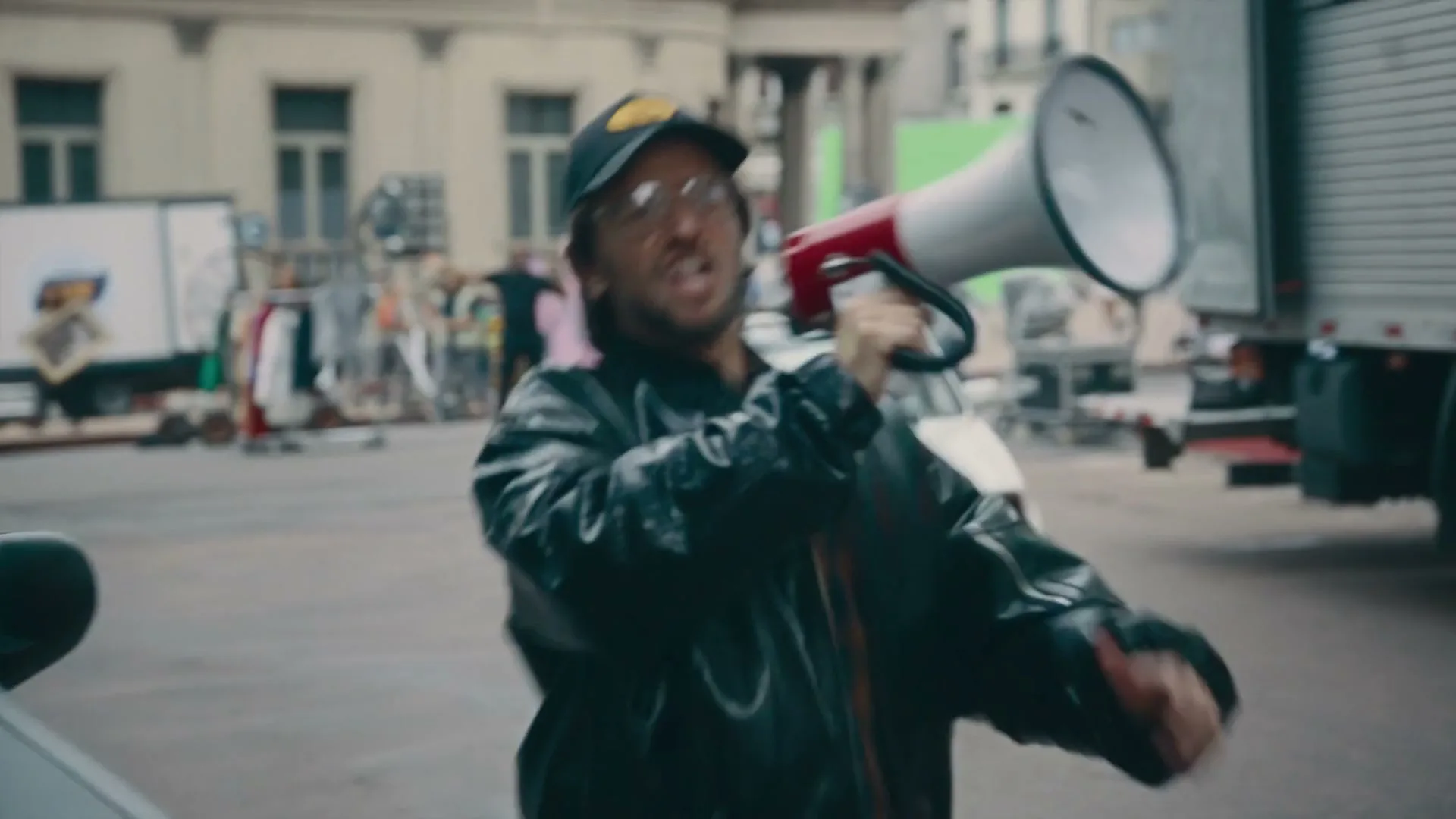 A male director in a black jacket and cap shouts into a megaphone on a busy city street, with cars and buildings in the background.