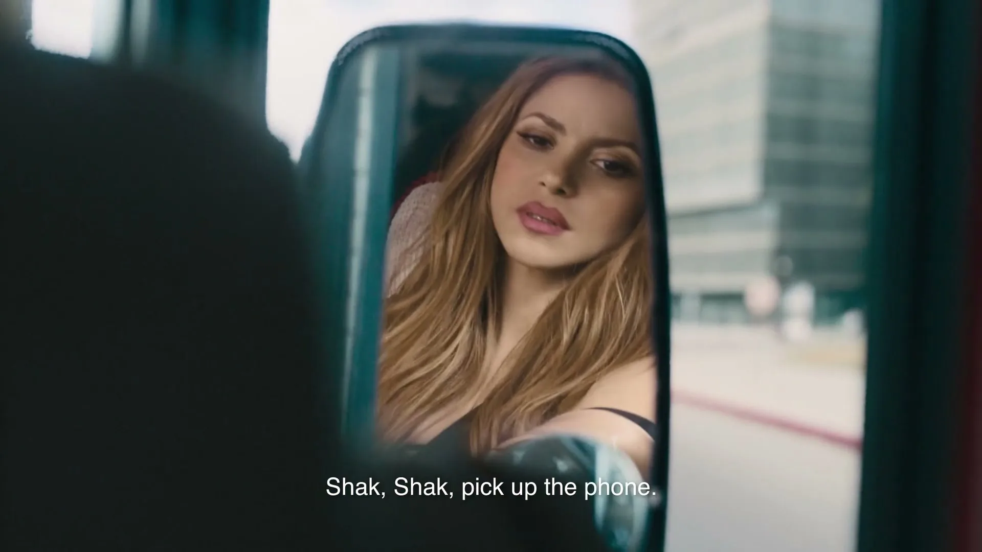 Shakira, a woman with long blonde hair, looks into a rearview mirror while sitting in a vehicle on a city street.