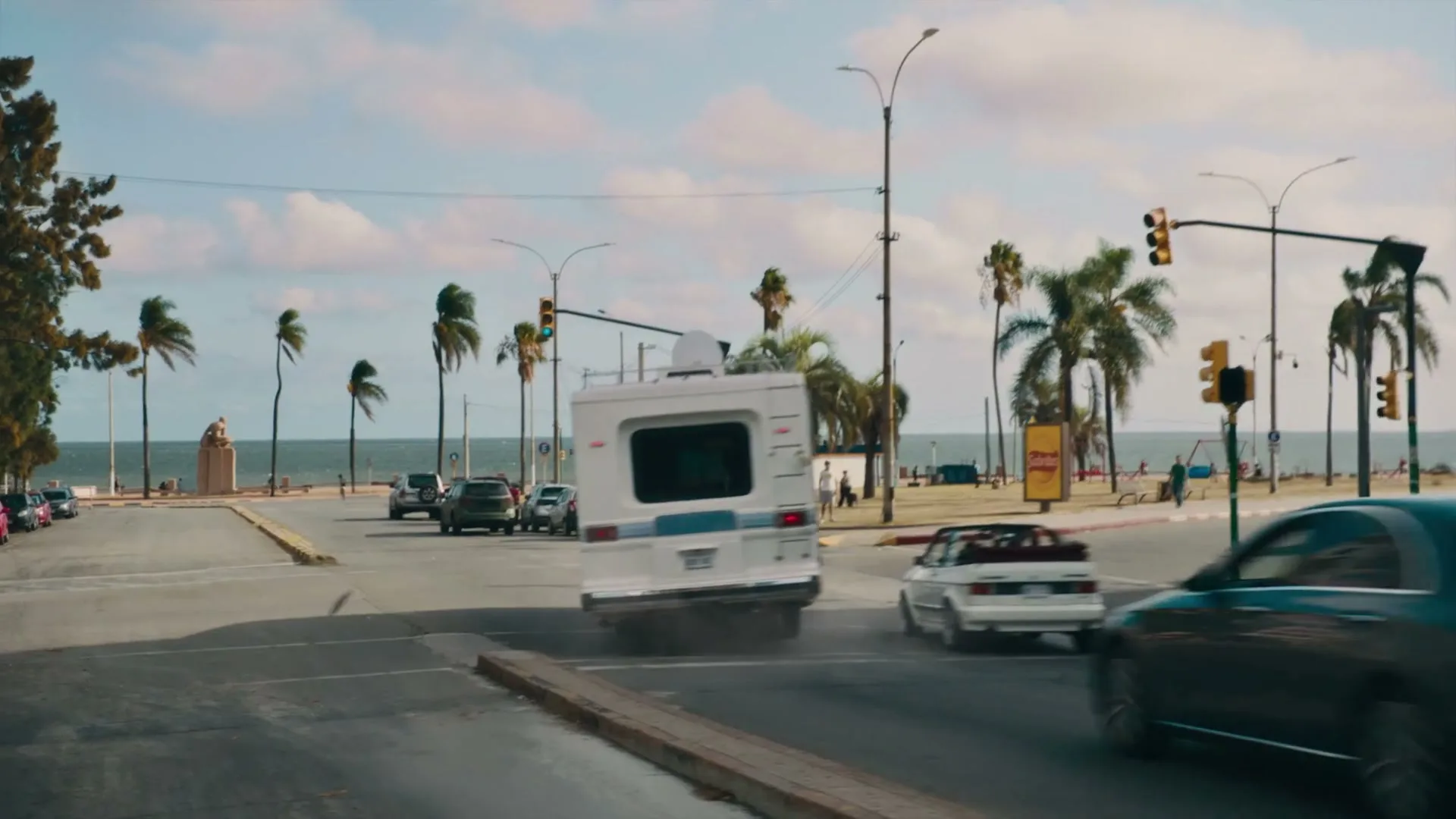 A white RV swerves sharply to the right, kicking up dust, as it turns a corner on a coastal road with palm trees and the ocean in the background.
