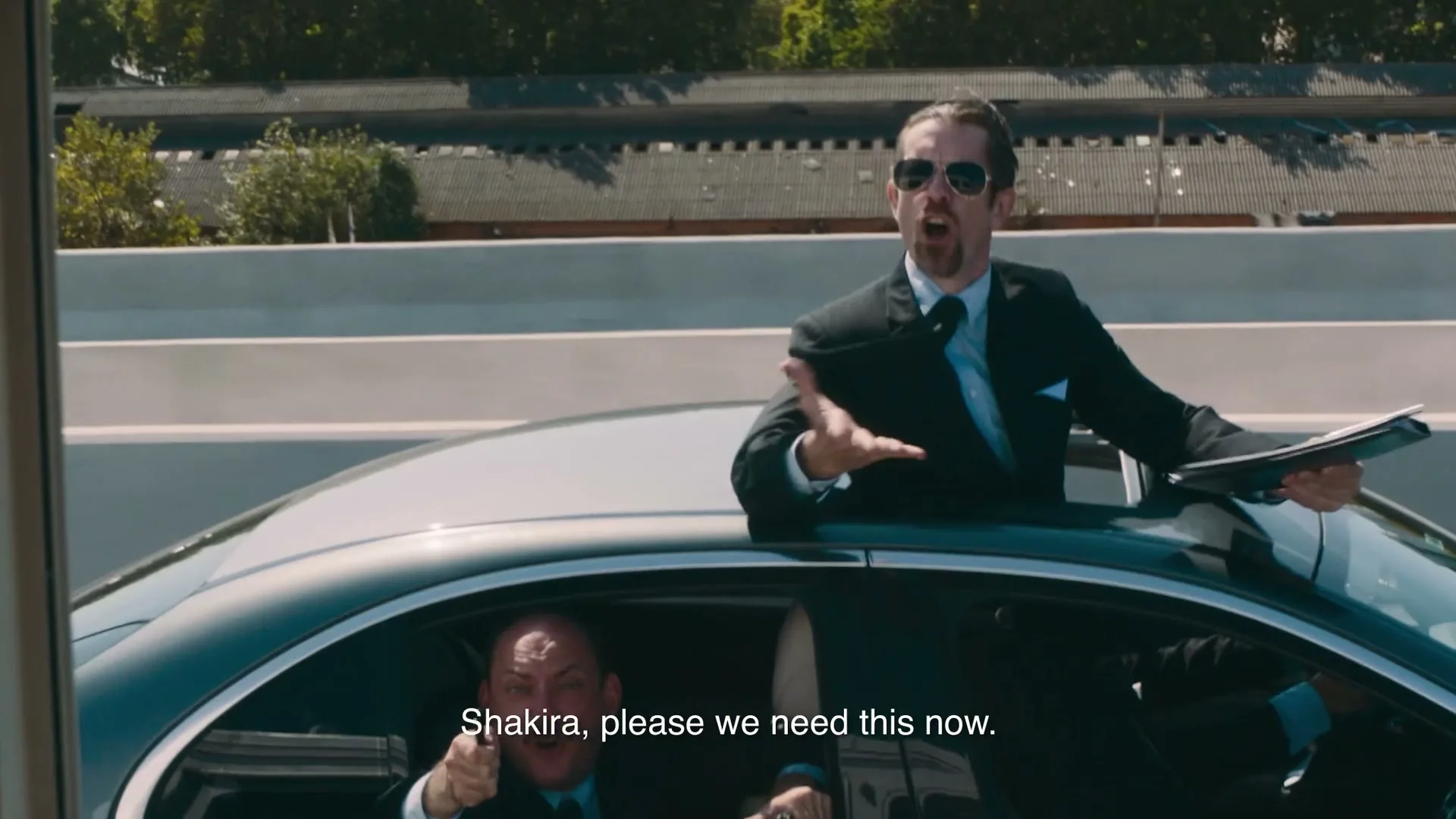 Two men in suits are in a car on a highway, one standing through the sunroof, both looking and gesturing urgently off-screen.