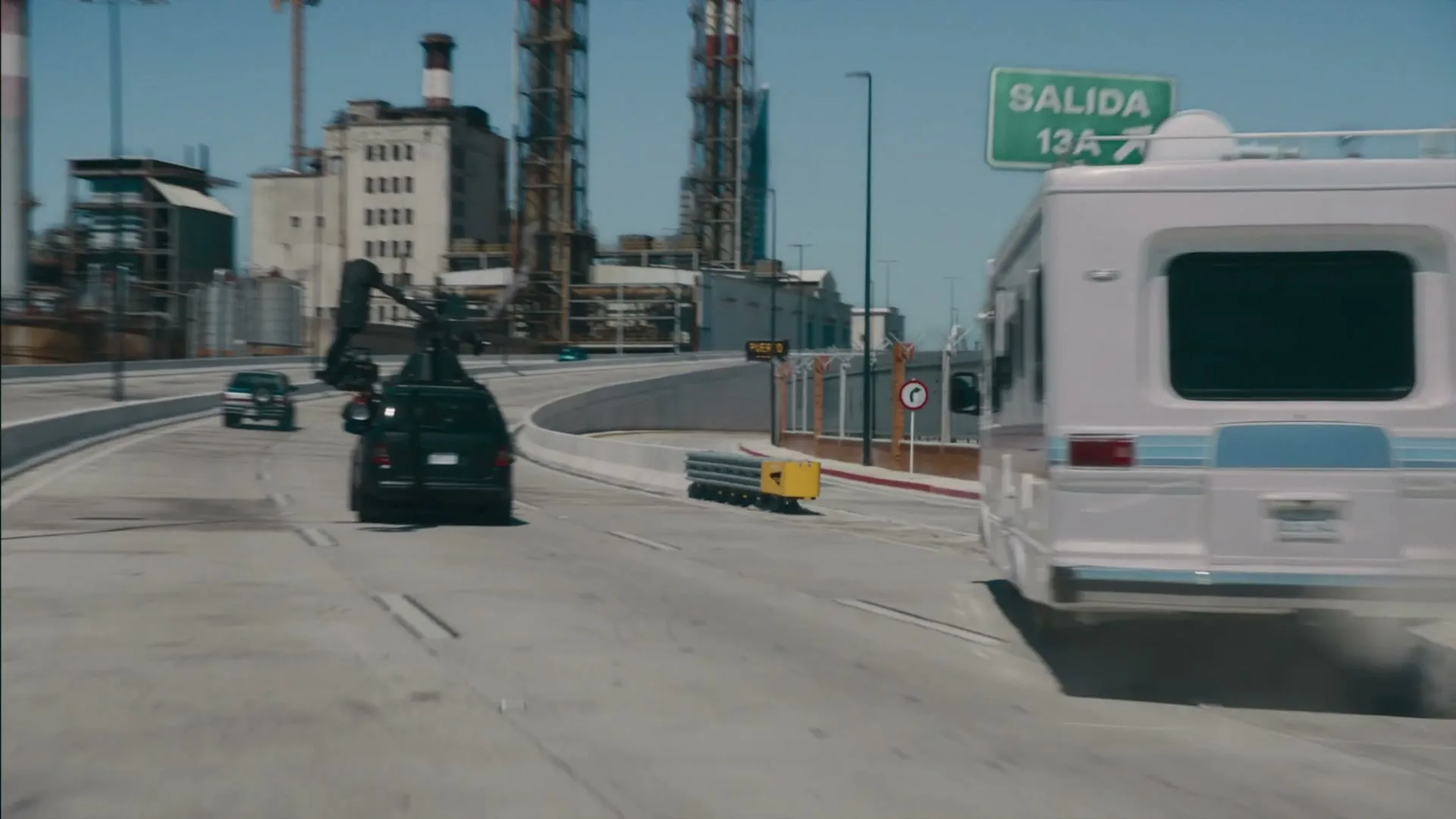 A white RV speeds down a highway, kicking up dust, with an industrial complex in the background under a clear sky.