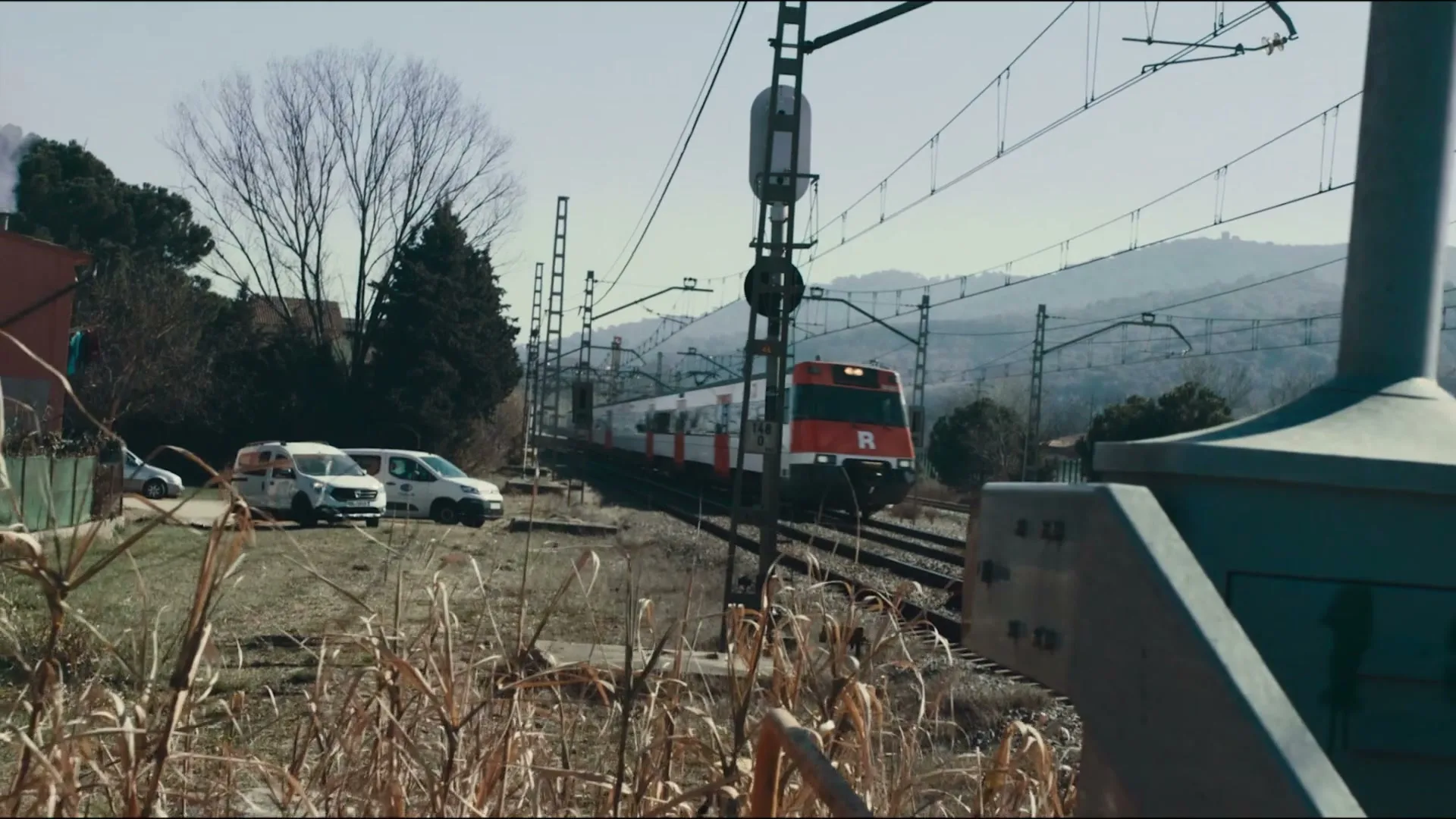 A red and white train travels along tracks through a rural landscape with mountains in the background and dry grass in the foreground.