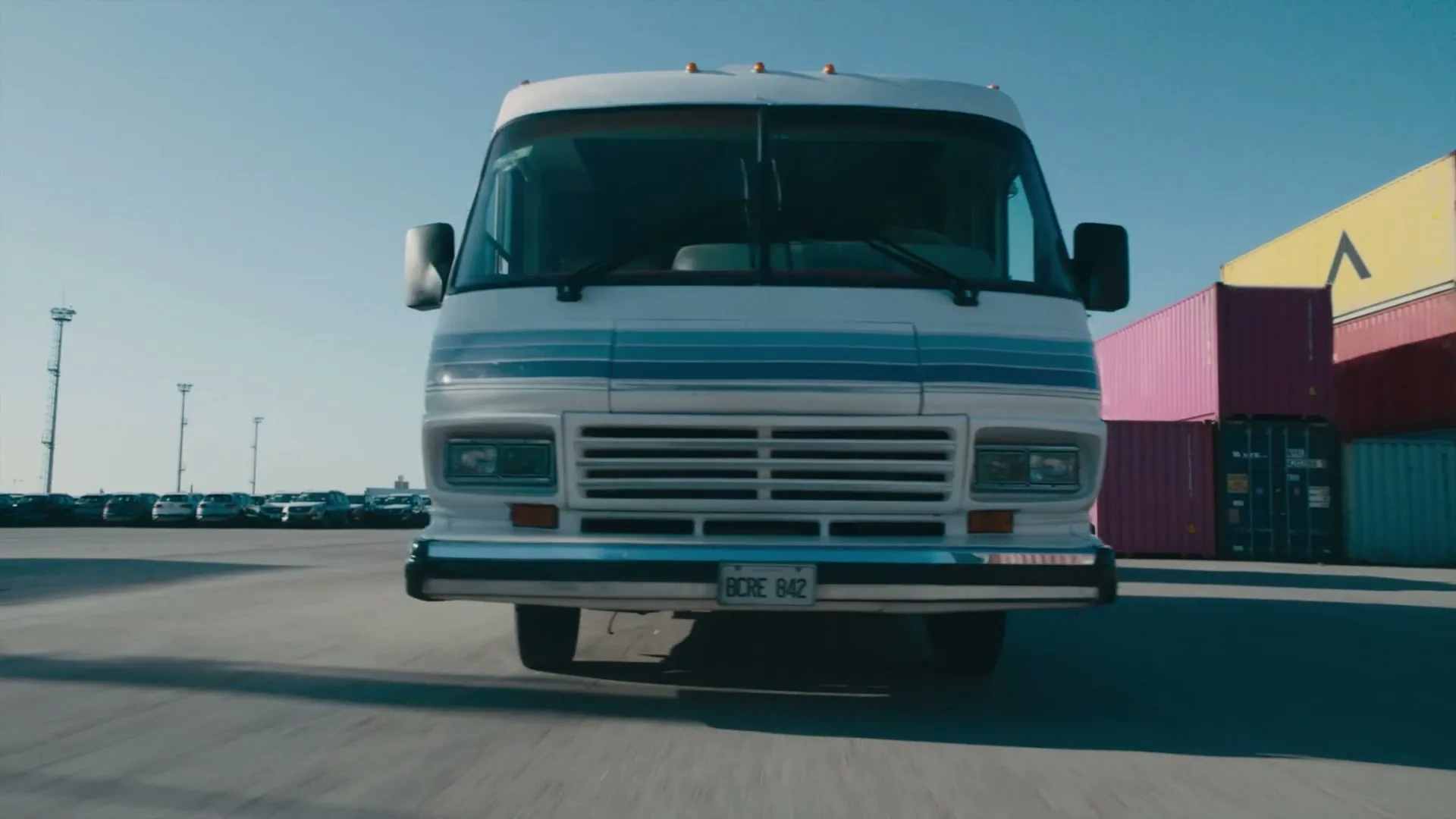 A white and blue RV drives towards the camera on a sunny day in a shipping yard with colorful containers in the background.