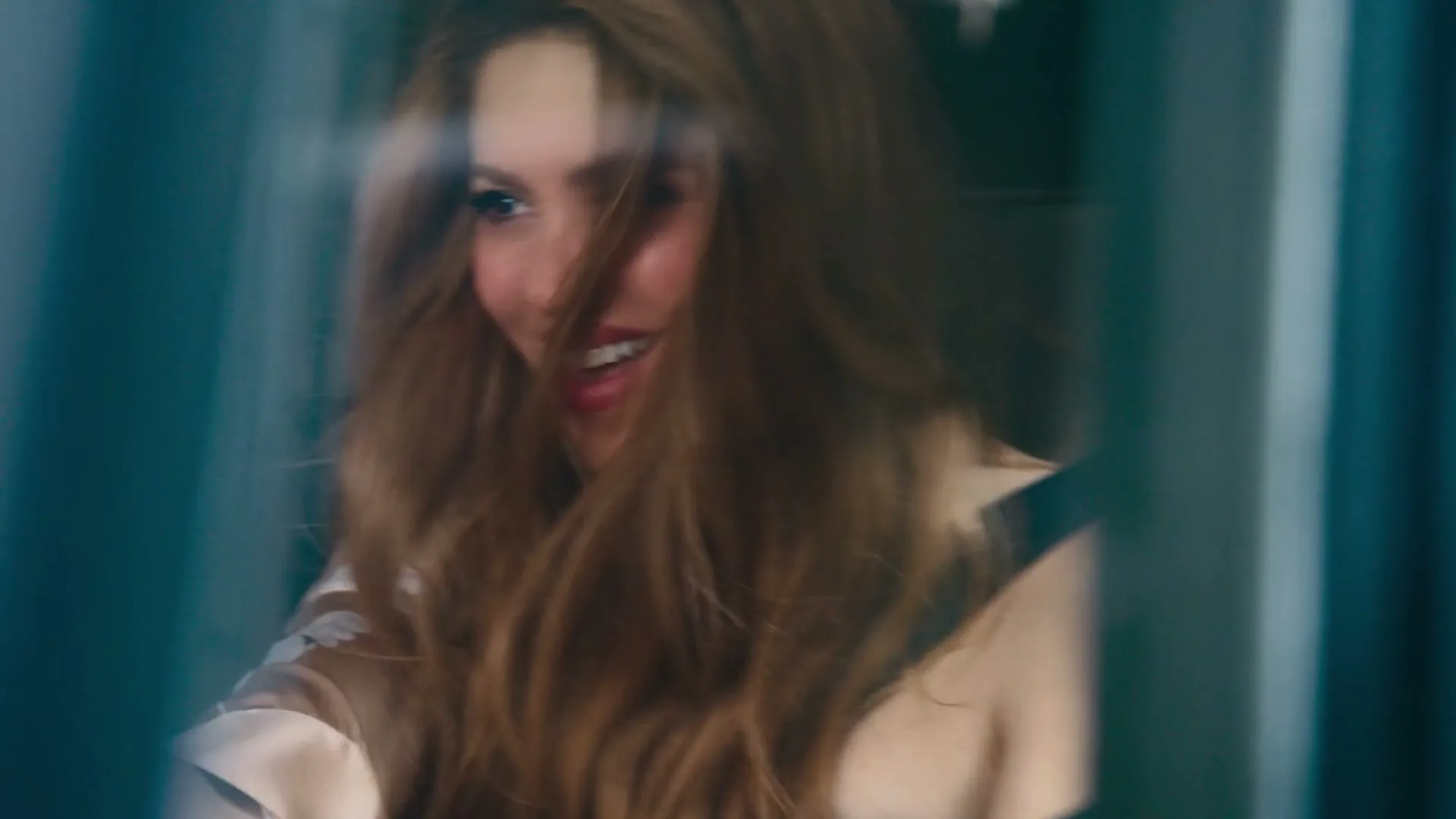 Shakira, a woman with long blonde hair, smiles and laughs while looking out a window on a train.
