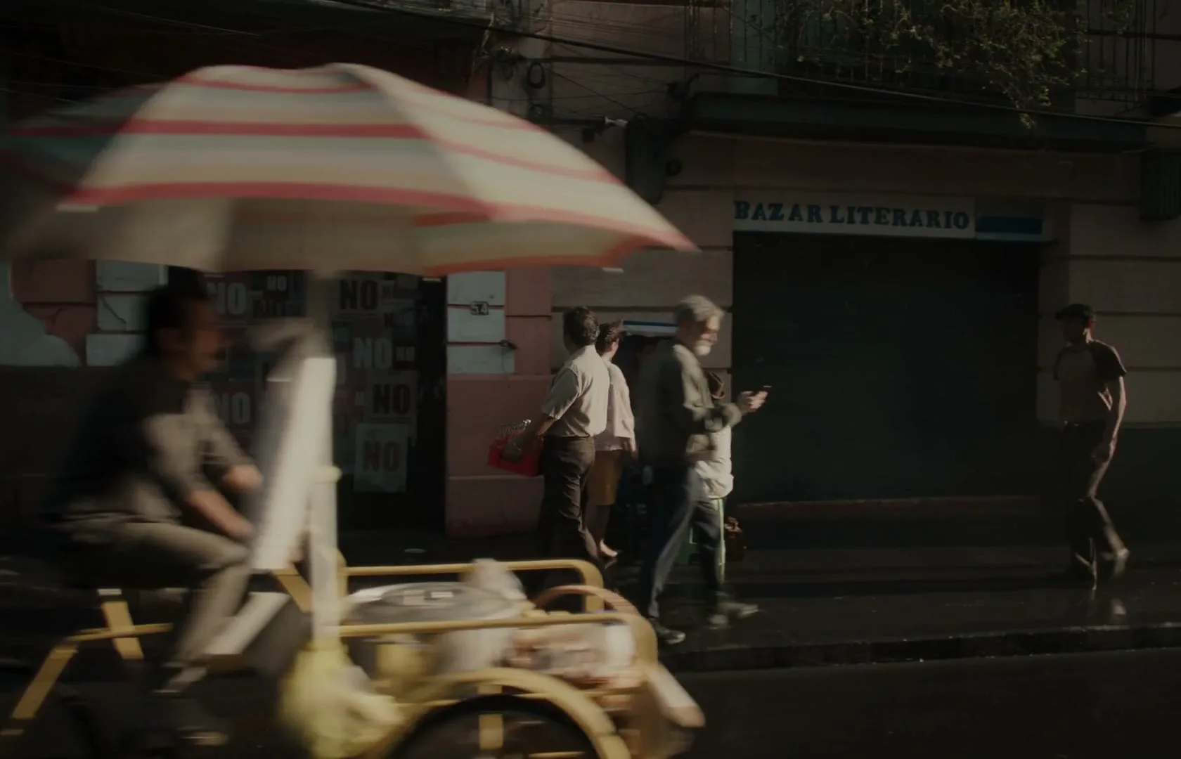 A diverse group of people walk and bike on a busy city street, passing a "Bazar Literario" and a wall covered in "NO" posters.