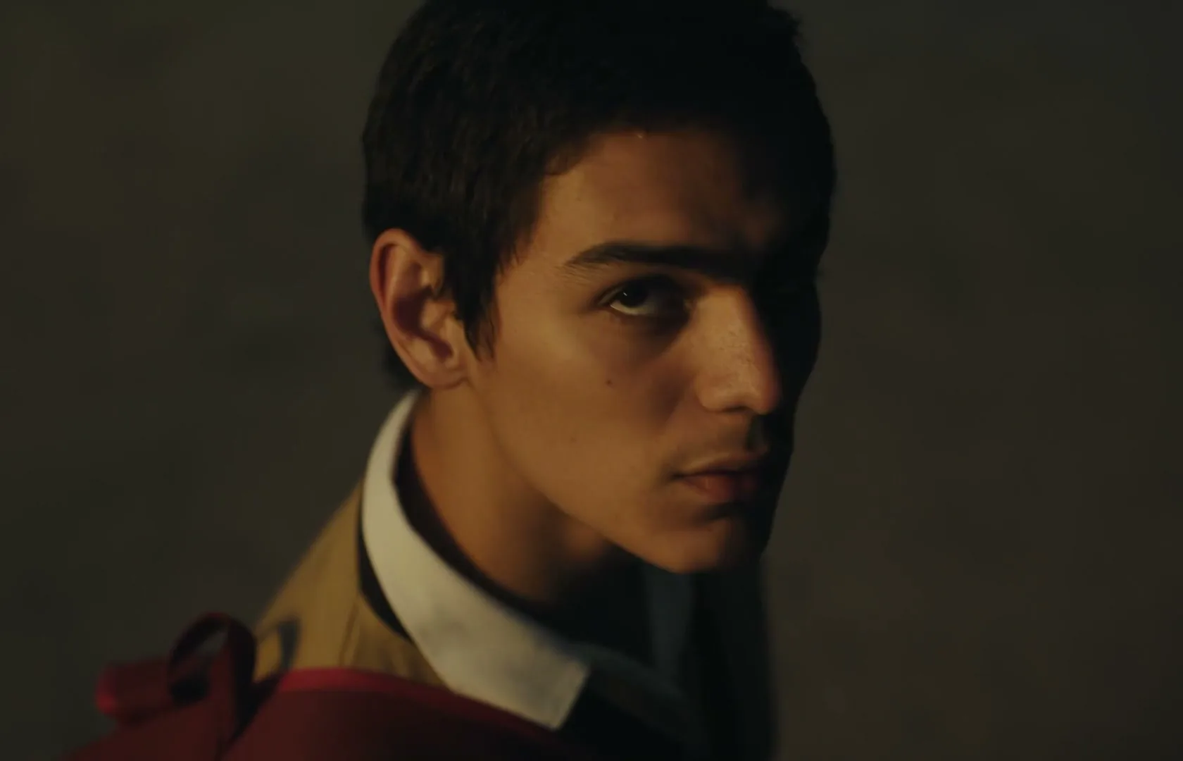 A young male with dark hair and a tan jacket turns his head to look at the camera in a dimly lit, dark room.