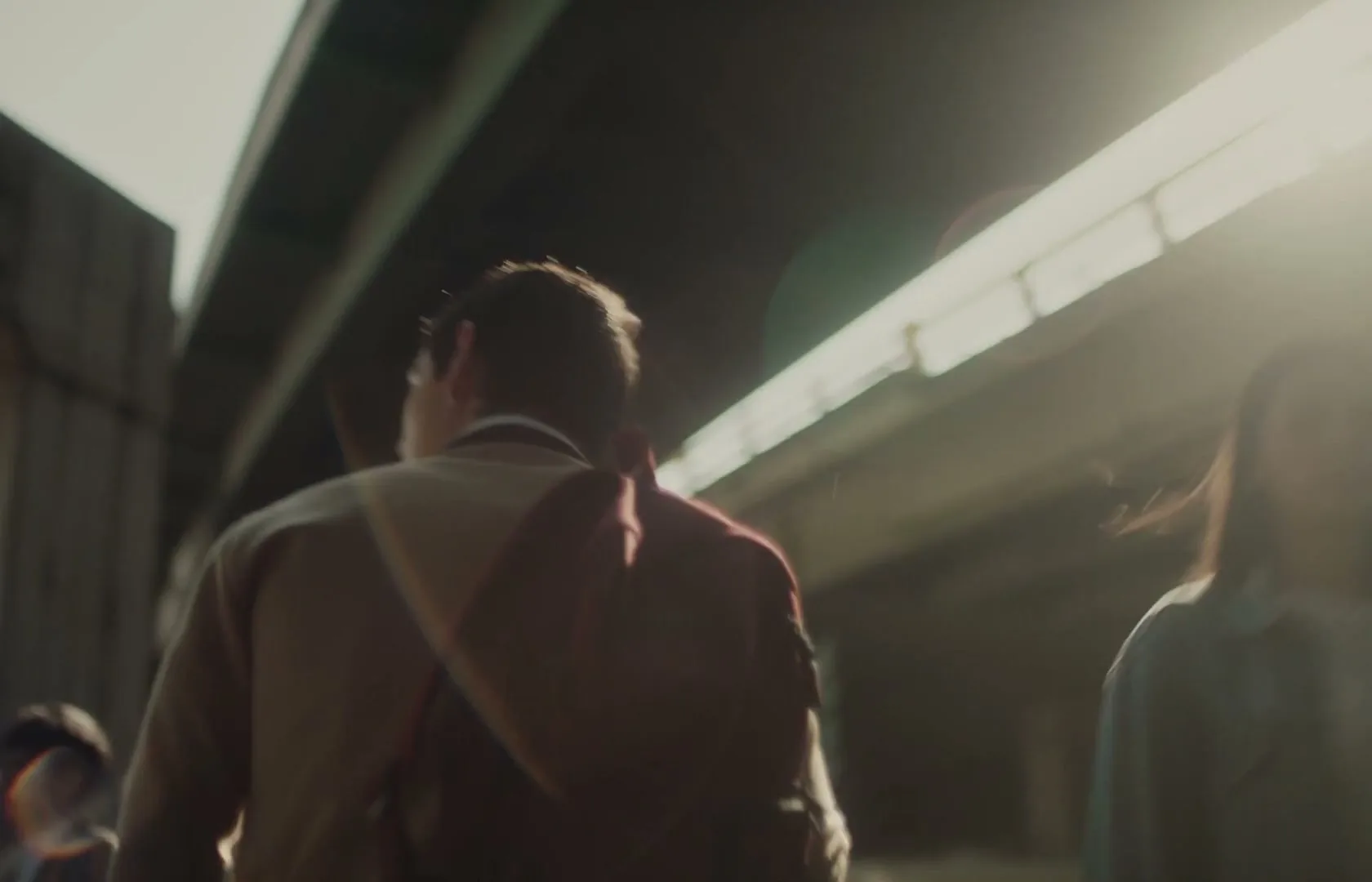 A young man with a backpack walks away from the camera under an overpass, with sunlight flaring from above.