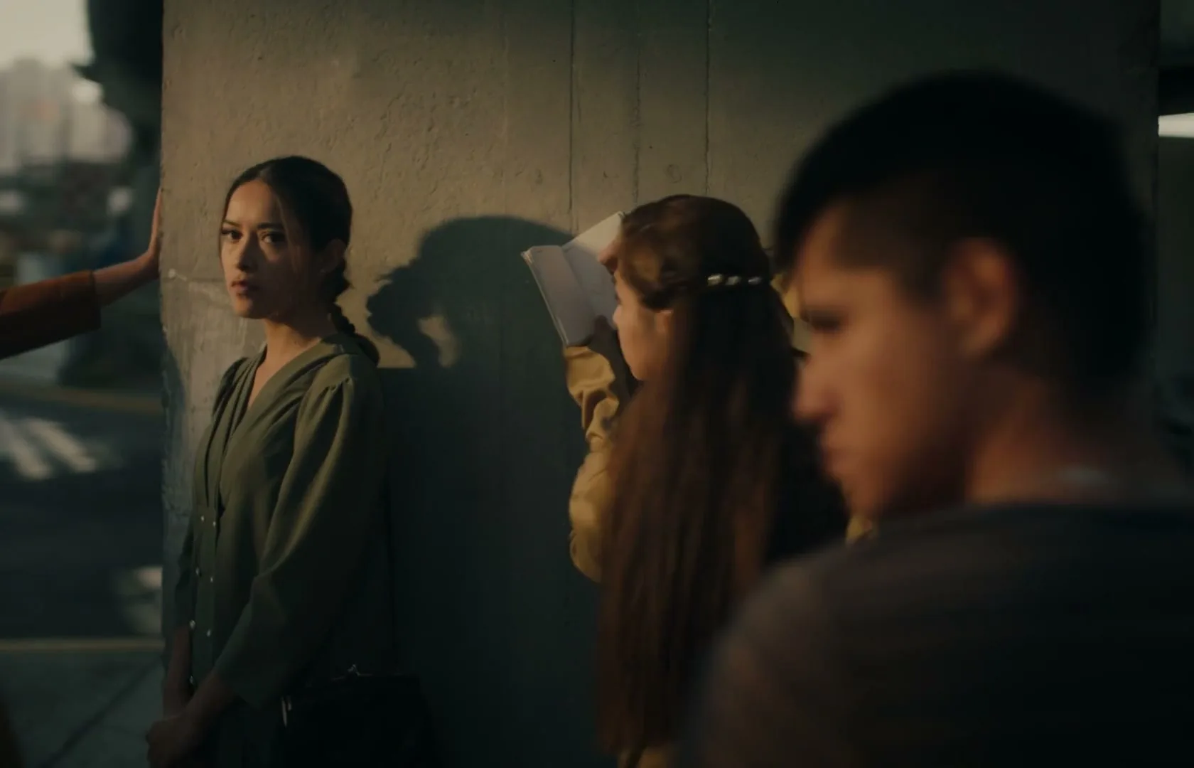 A young woman with a braid leans against a concrete wall, looking off-camera, while another young woman writes in a notebook next to her.