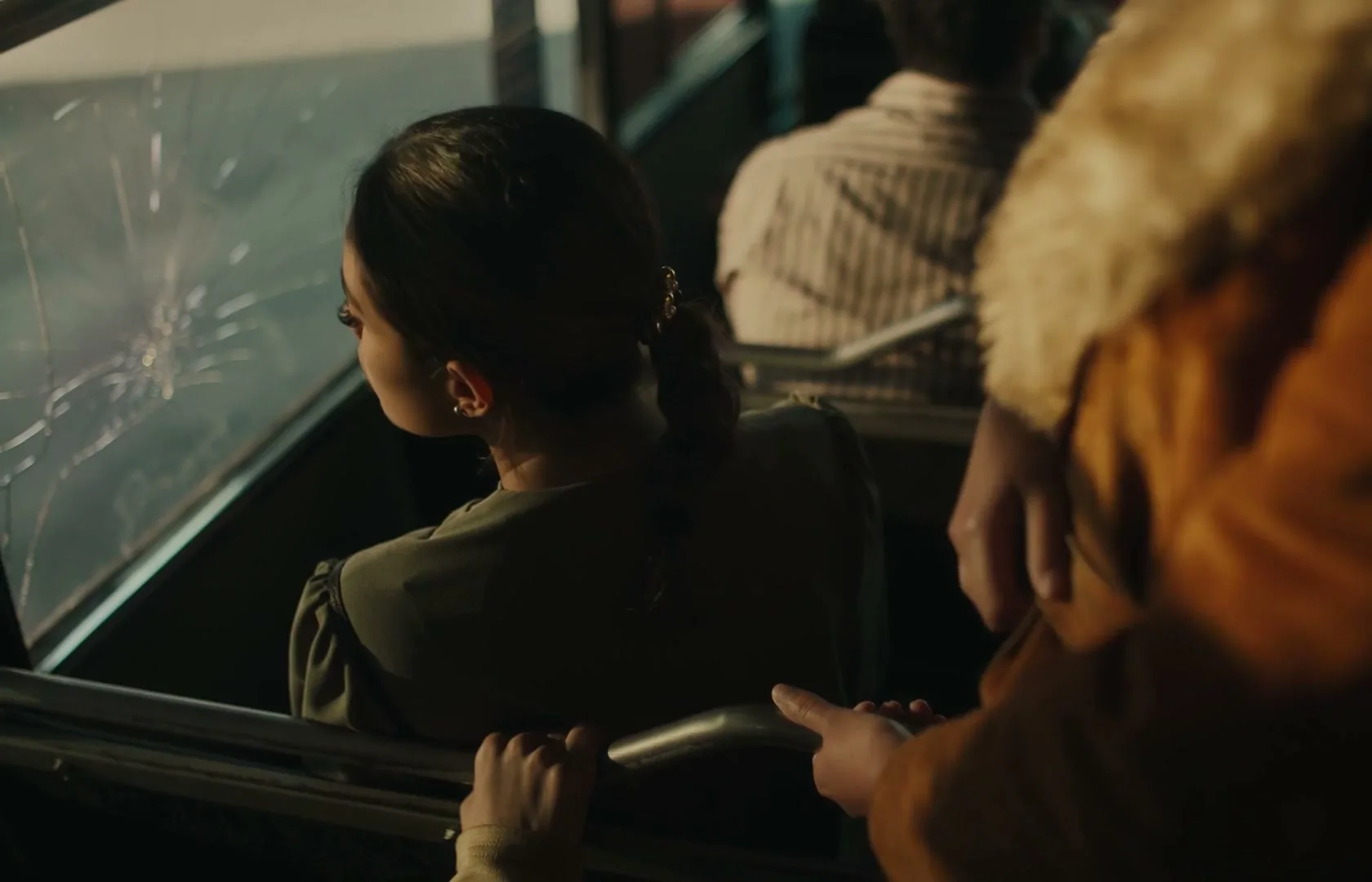 A young woman with a braided ponytail looks out a cracked bus window, while another person's arm is visible in the foreground.