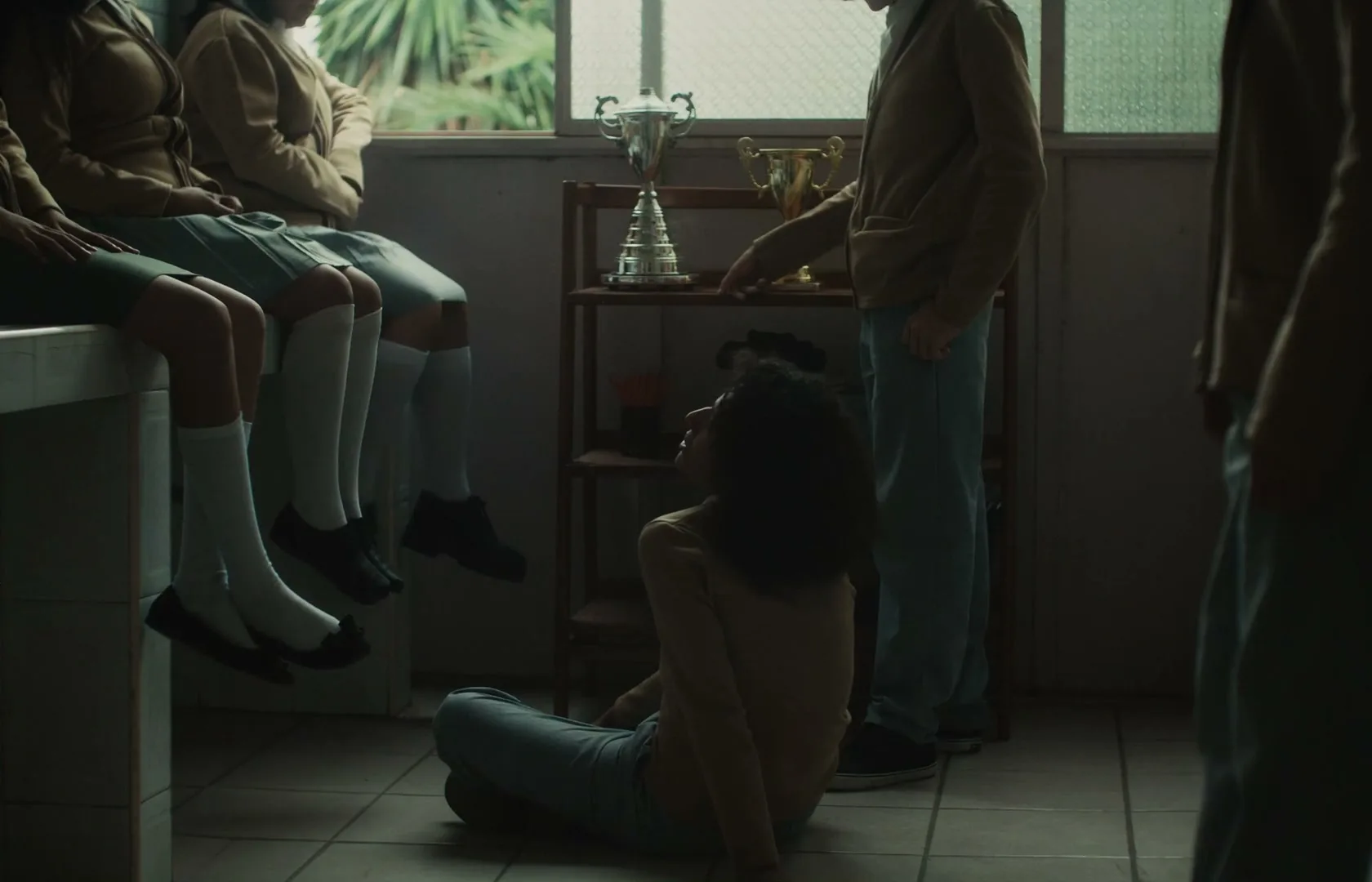 A group of high school students in uniform are gathered in a dimly lit room, with some sitting on a counter and one on the floor, looking up at a boy standing near a shelf with trophies.