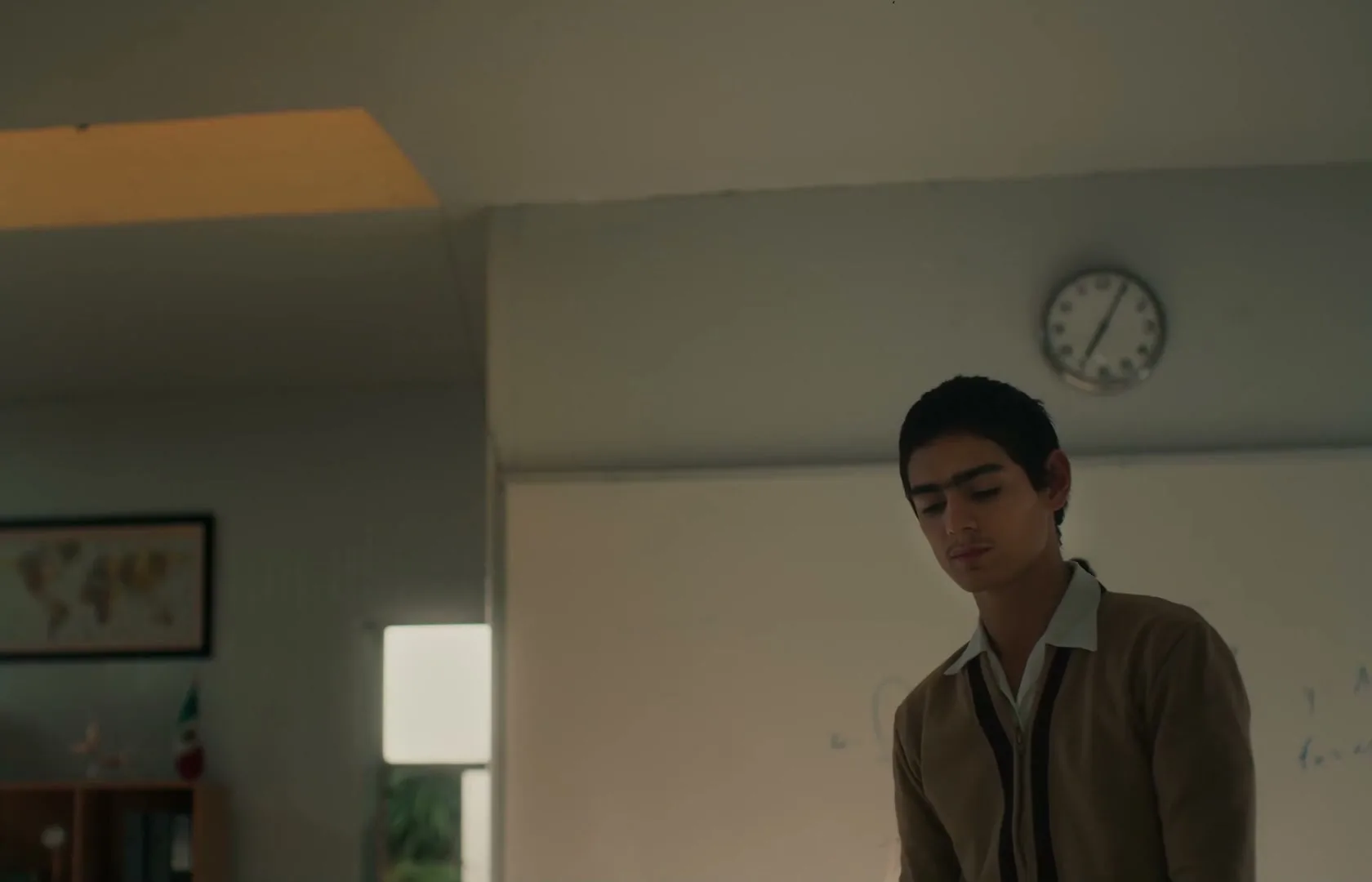 A male teen in a brown cardigan looks down sadly in a classroom, while a female teen walks past him.