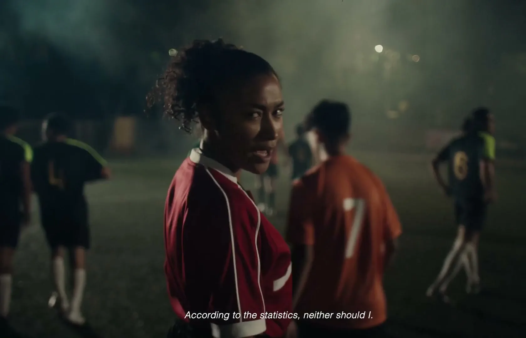 A young female soccer player in a red jersey looks back at the camera with a determined expression on a dimly lit field at night, surrounded by other players.