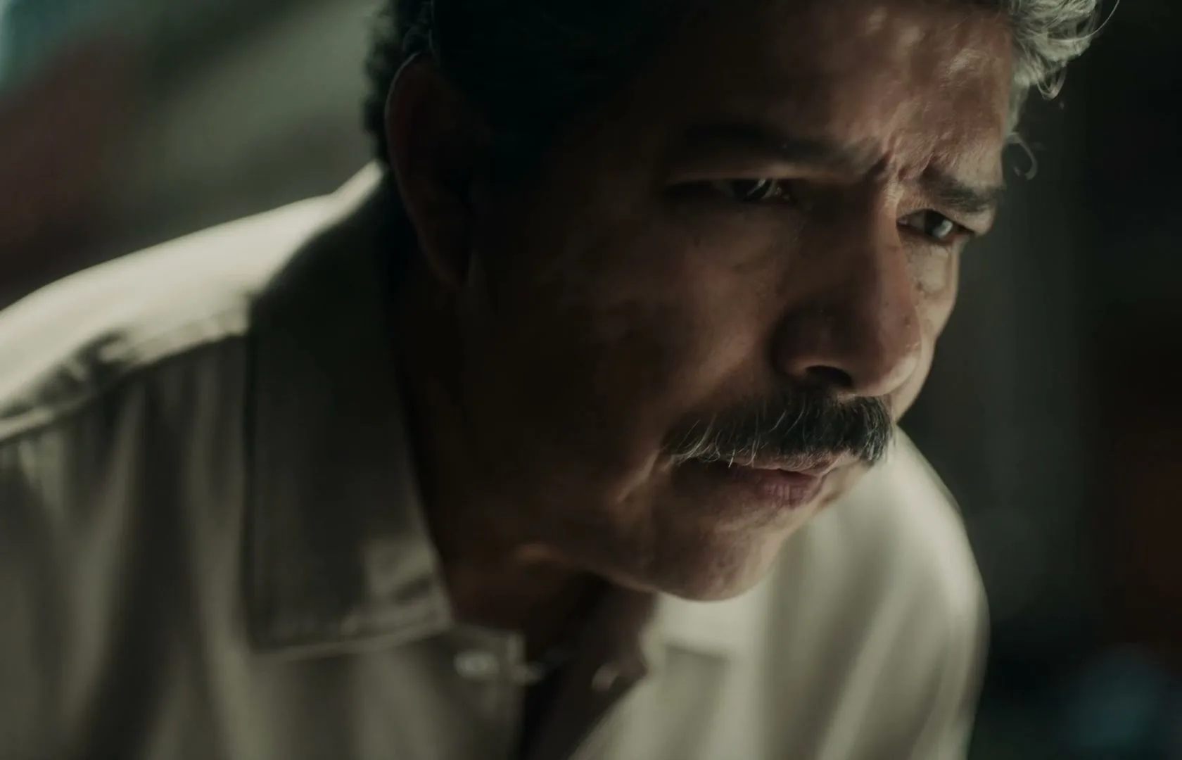 A middle-aged man with a mustache and graying hair looks down with a concerned expression, illuminated by soft light in an interior setting.