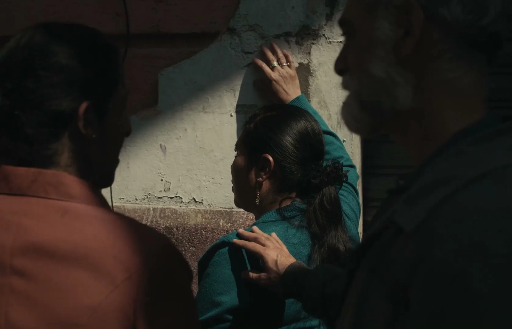 A woman in a teal sweater leans against a wall, comforted by a man's hand on her shoulder, while another person stands nearby.