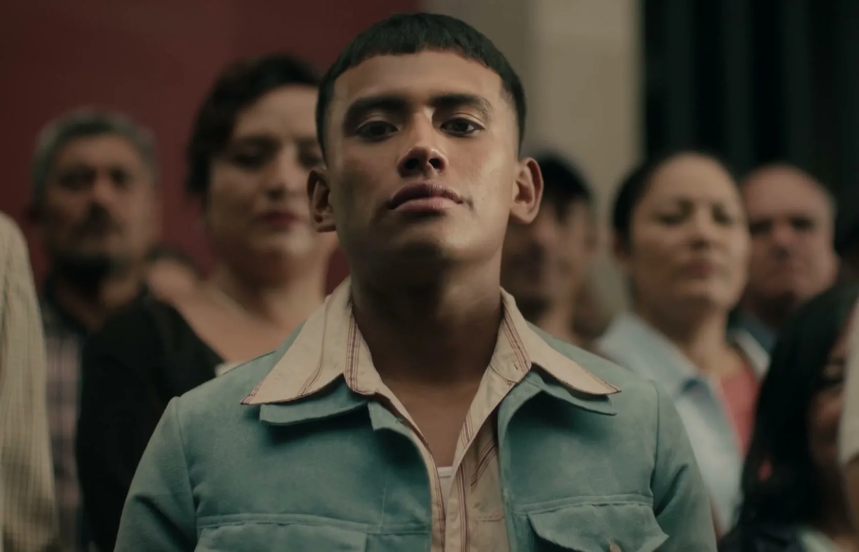 A young man with a short haircut and a light blue jacket stands confidently, looking directly at the camera amidst a blurred crowd of people.