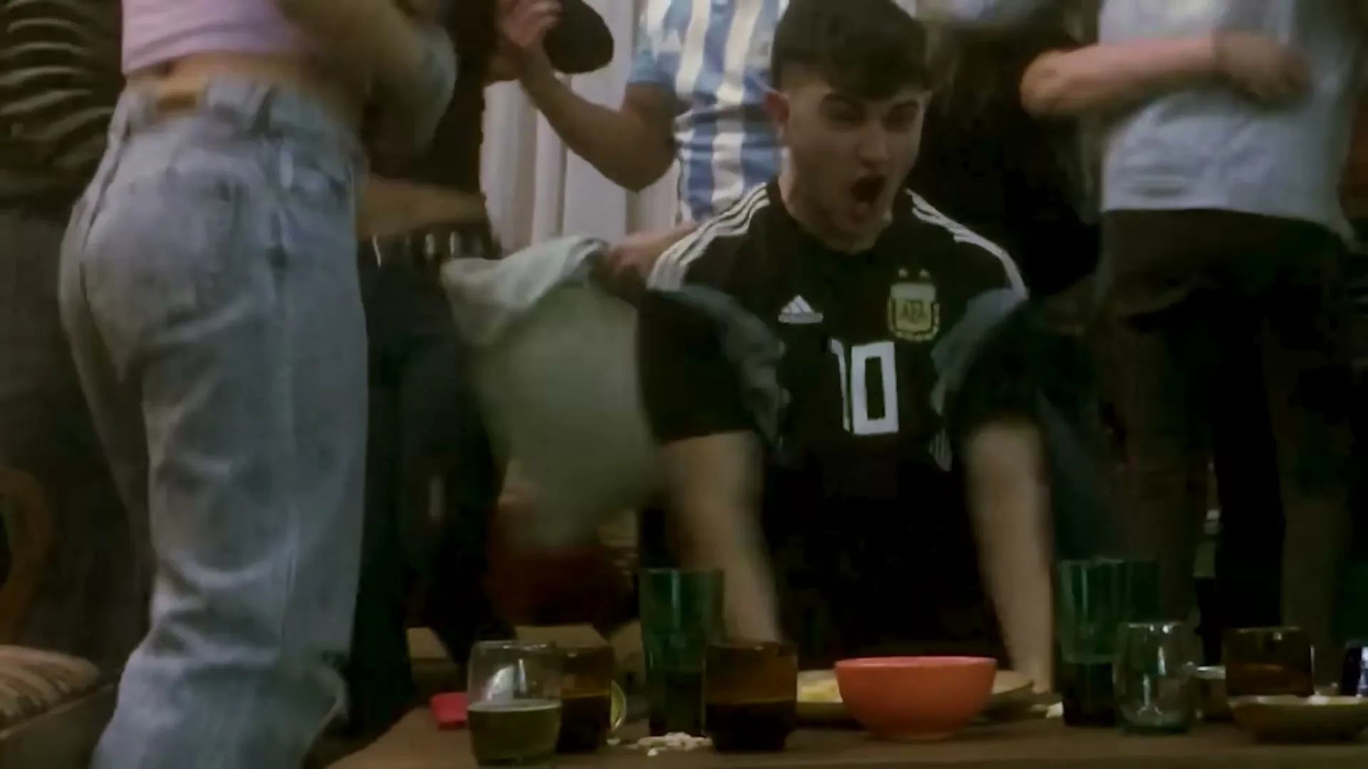 A group of young adults, wearing Argentina soccer jerseys, watch a game intently on a couch, then erupt in cheers and hugs when their team scores.