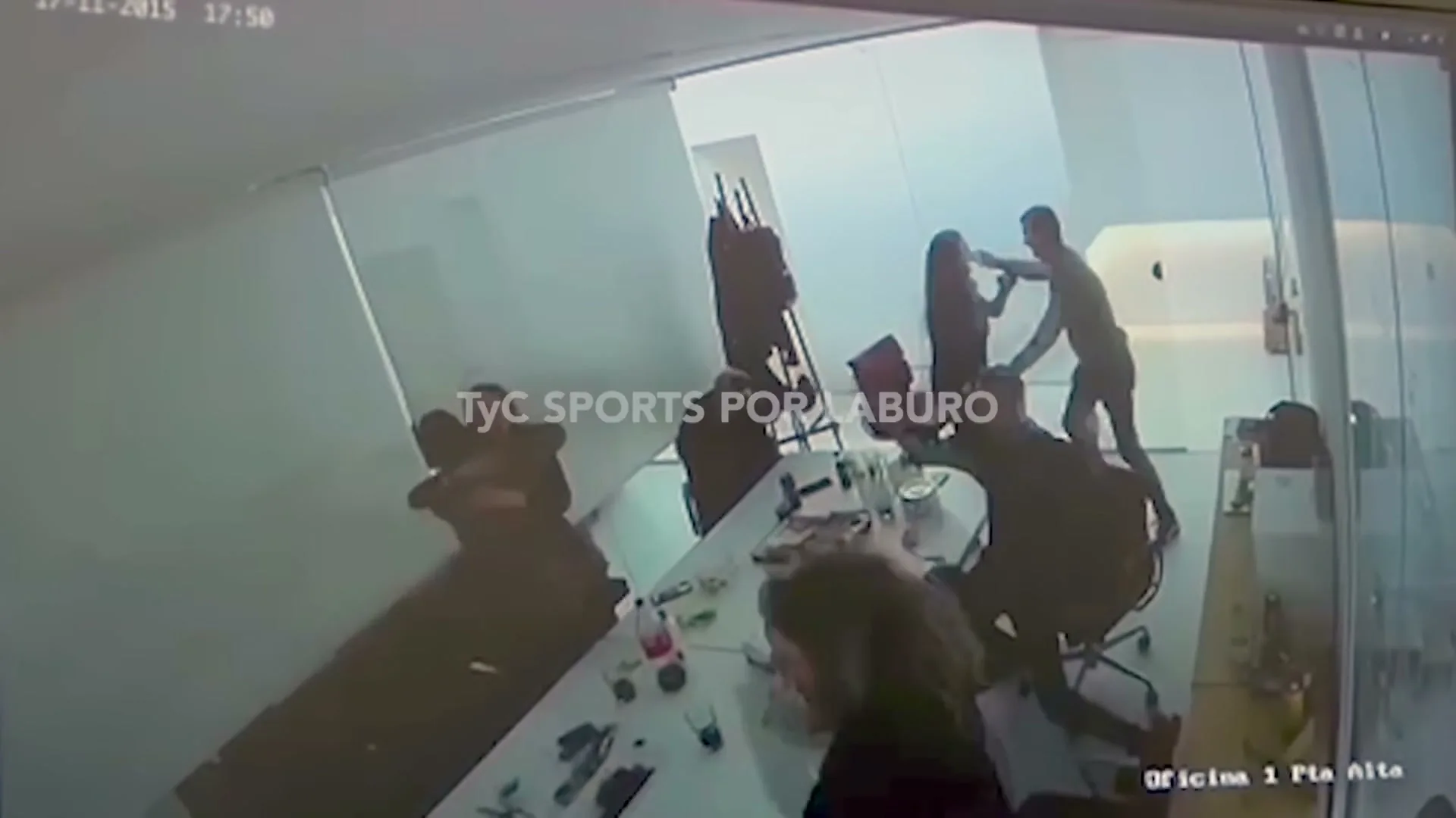 A group of office workers in dark clothing are seated around a conference table, then jump up and cheer excitedly in an office.