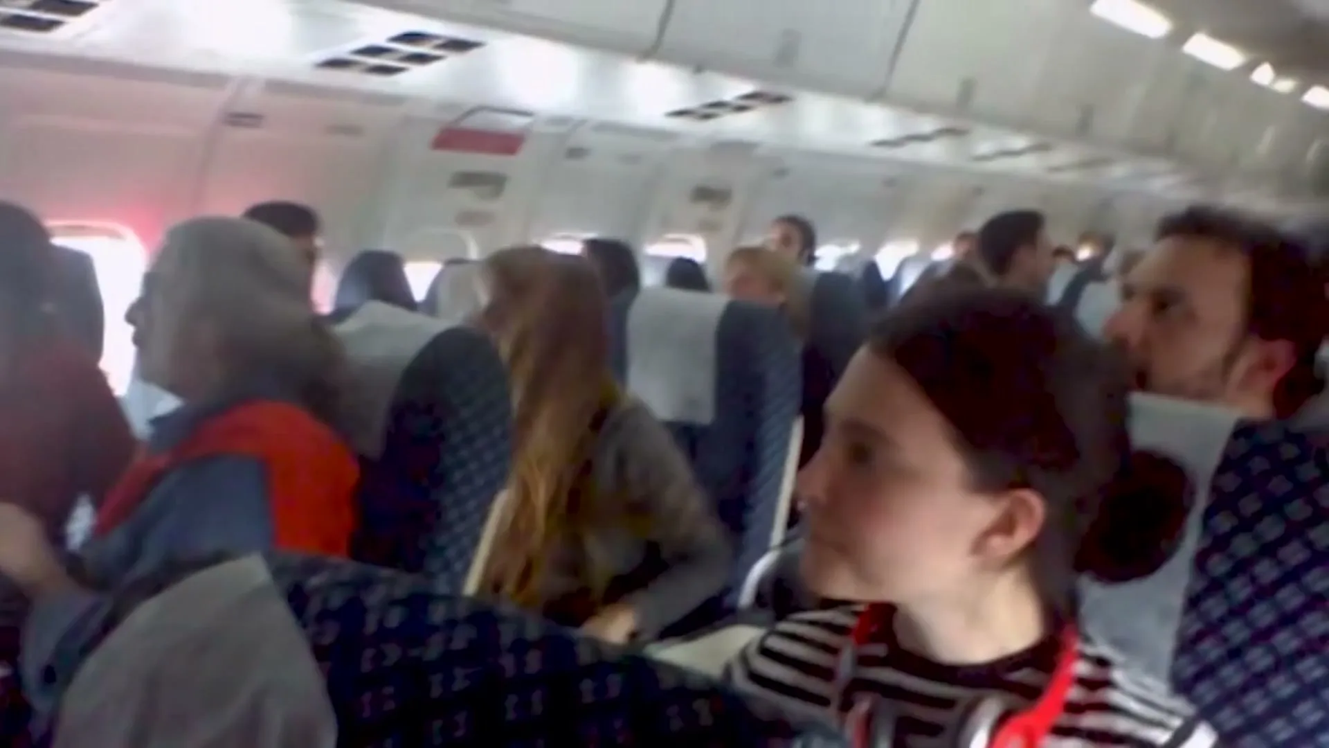 Passengers on an airplane react with surprise and excitement as they hear news of a goal, turning their heads and opening their mouths in disbelief.