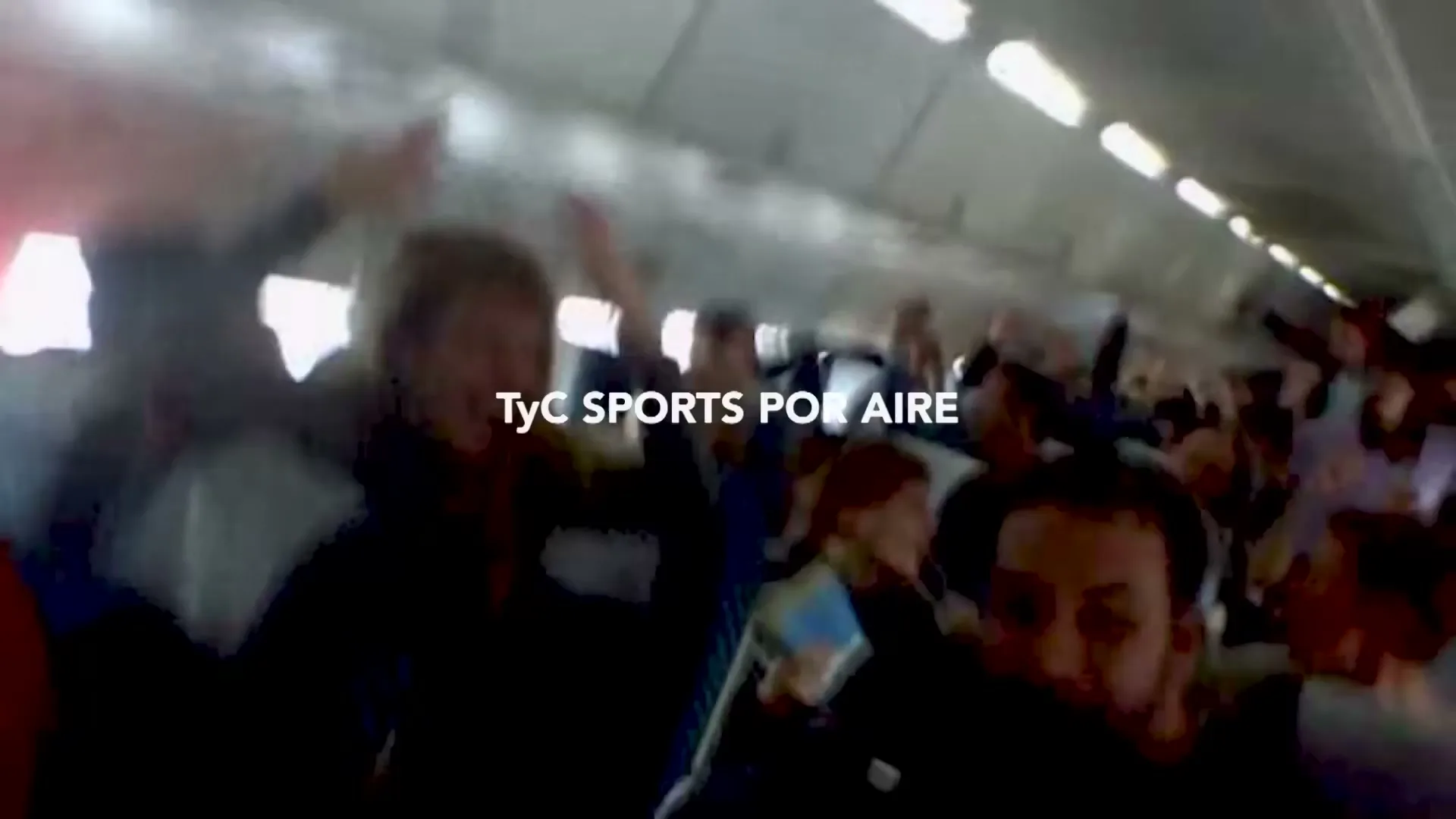 A crowd of people, some with their arms raised, are seen in a blurry, shaky shot inside what appears to be an airplane cabin.