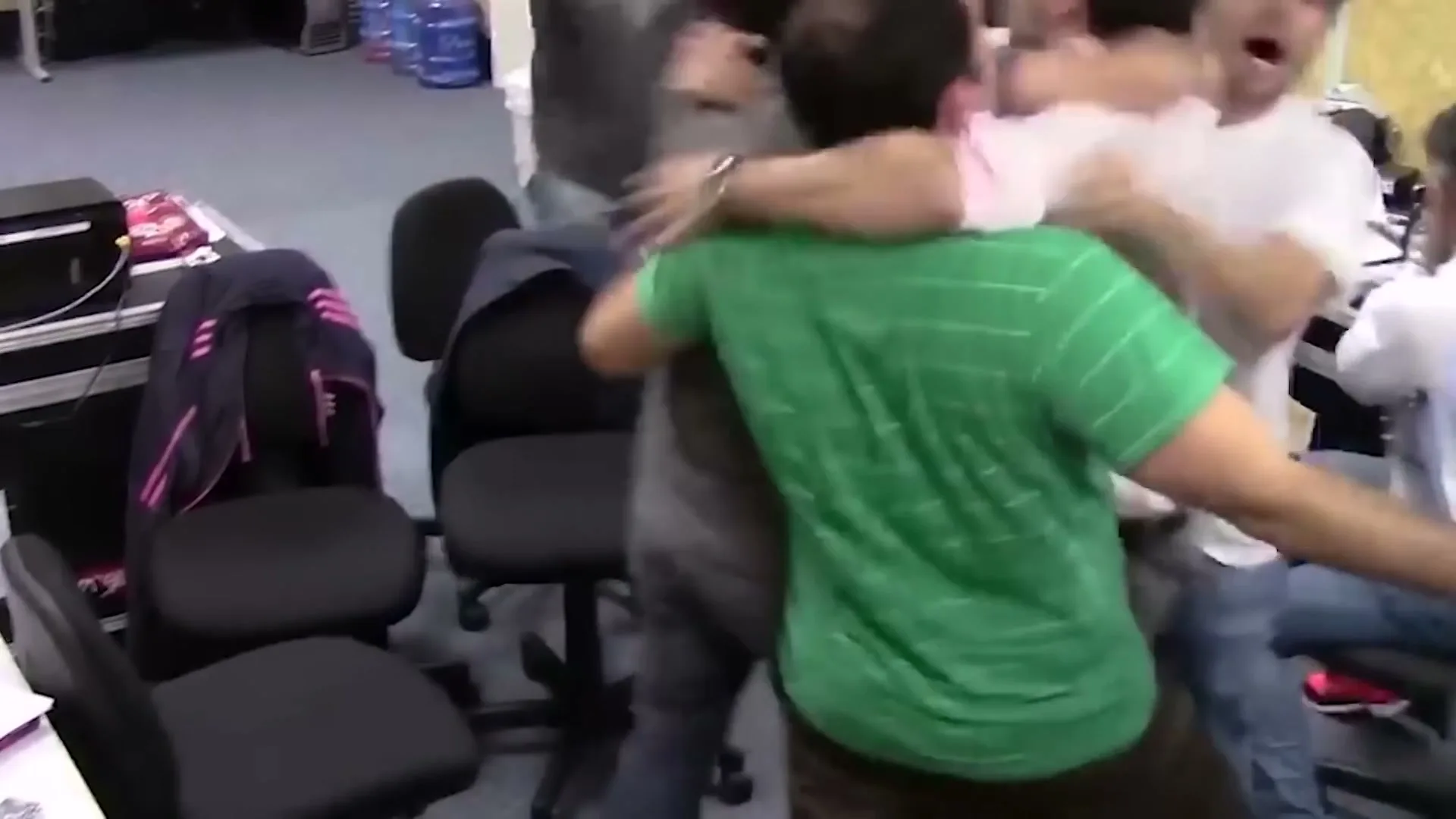 A group of men in an office setting jump up and embrace each other in celebration, with one man in a green striped shirt prominently in the foreground.