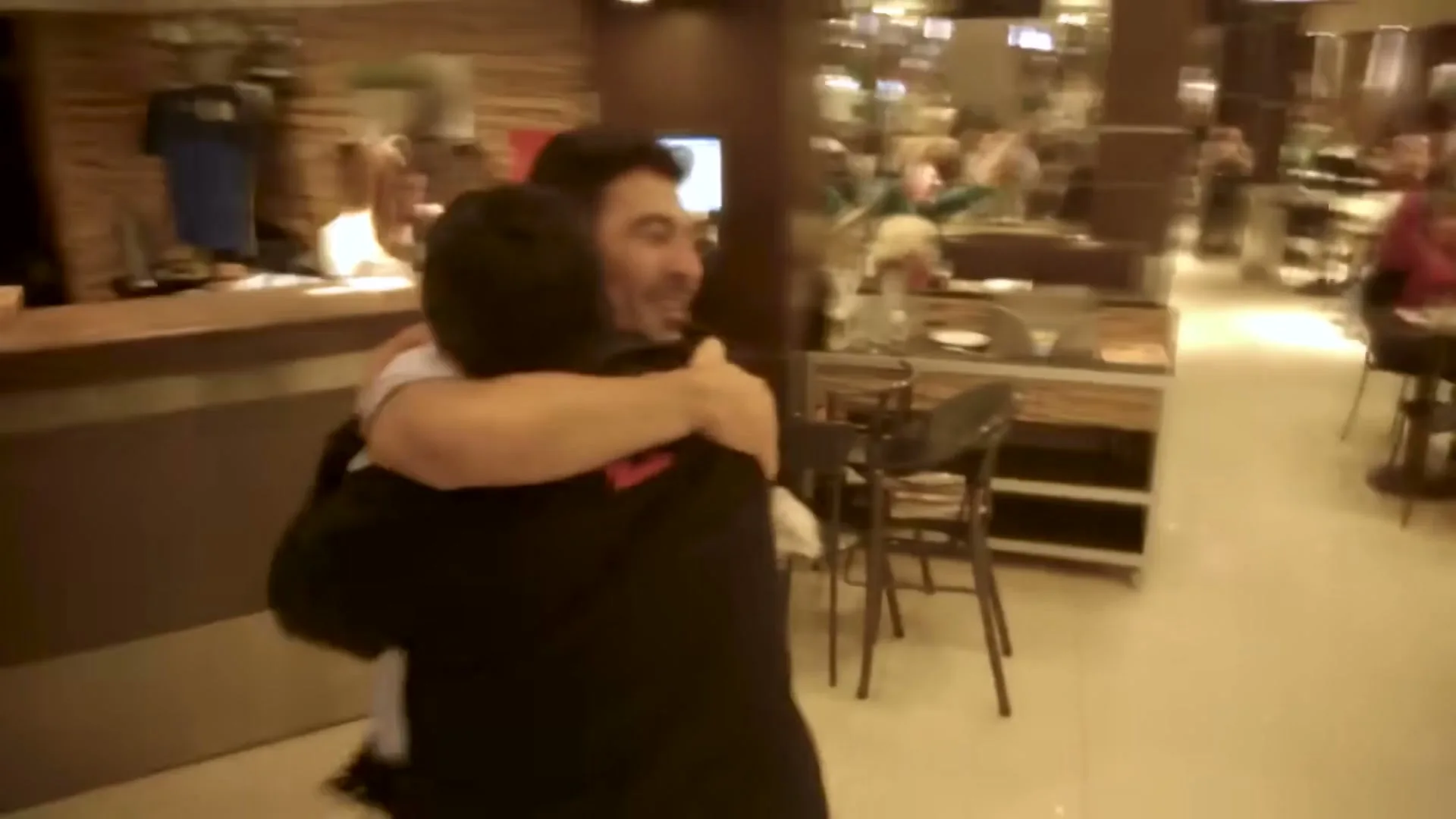 Two men embrace in a joyful hug in a restaurant, with one man looking surprised and happy, surrounded by other patrons.