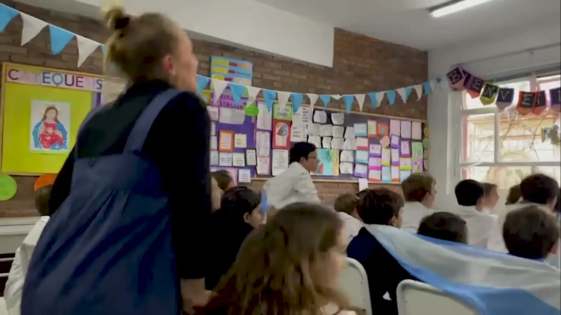 A female teacher in a blue apron stands in a classroom, looking towards a window as students in white shirts turn their heads, some with an Argentinian flag.