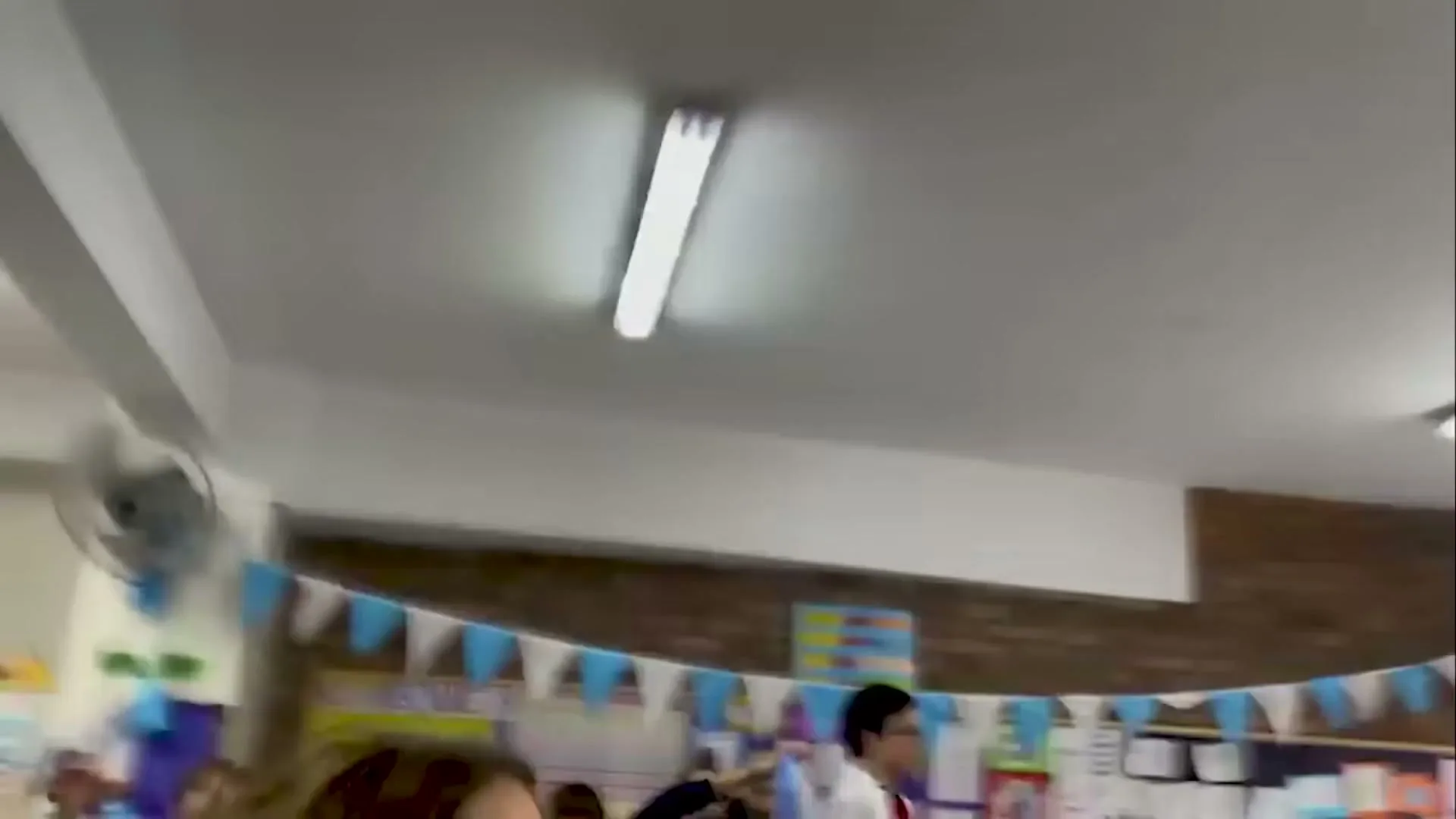 A blurry, fast-moving shot of a classroom with children and adults, decorated with blue and white bunting, as text appears on screen.