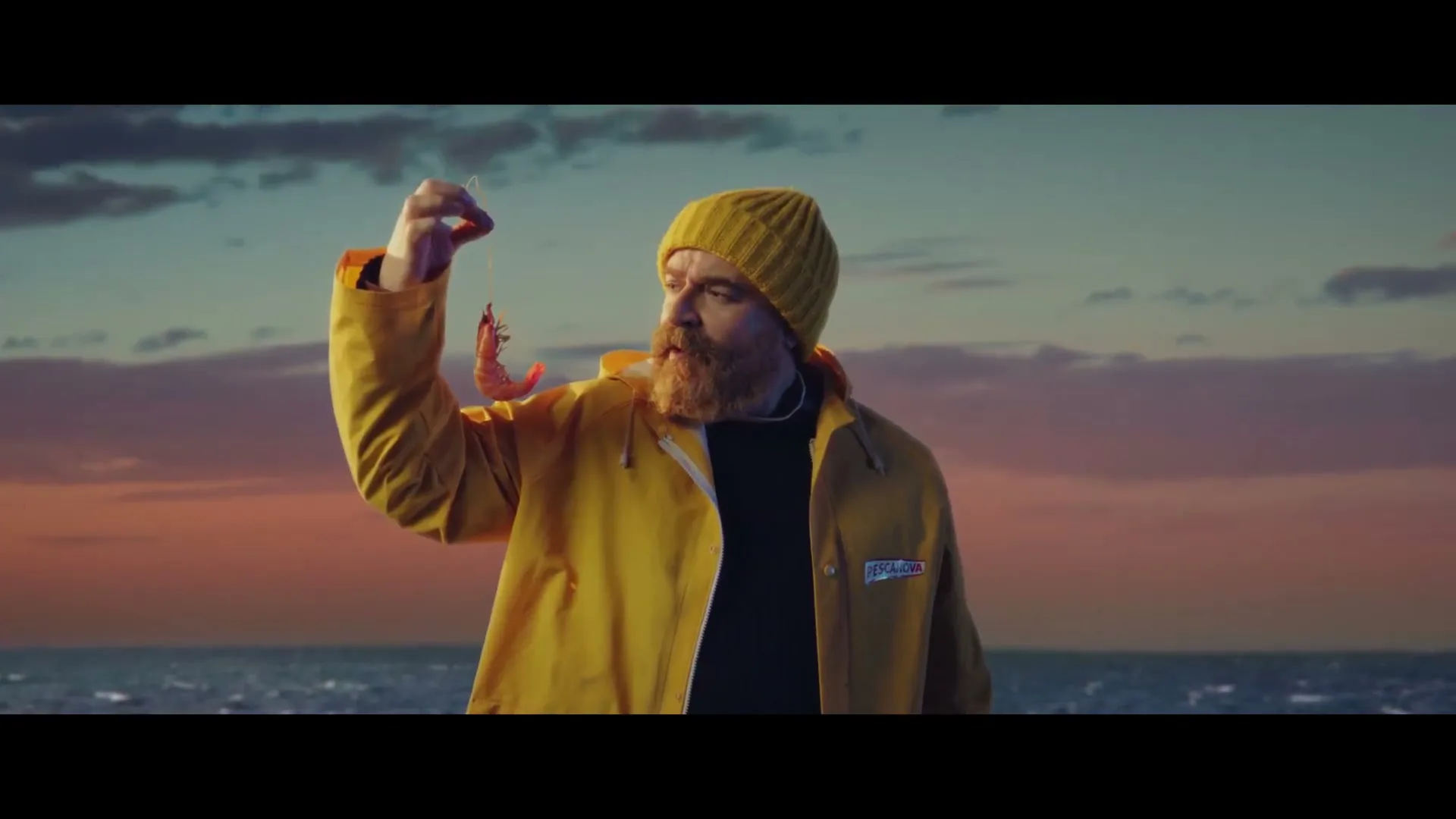 A bearded man in a yellow raincoat and beanie holds up a shrimp, looking at it with a thoughtful expression against a sunset ocean backdrop.