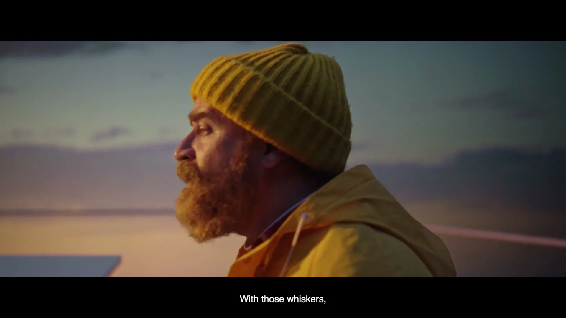 A bearded man in a yellow beanie and jacket looks out over the water at sunset, with a serene expression.