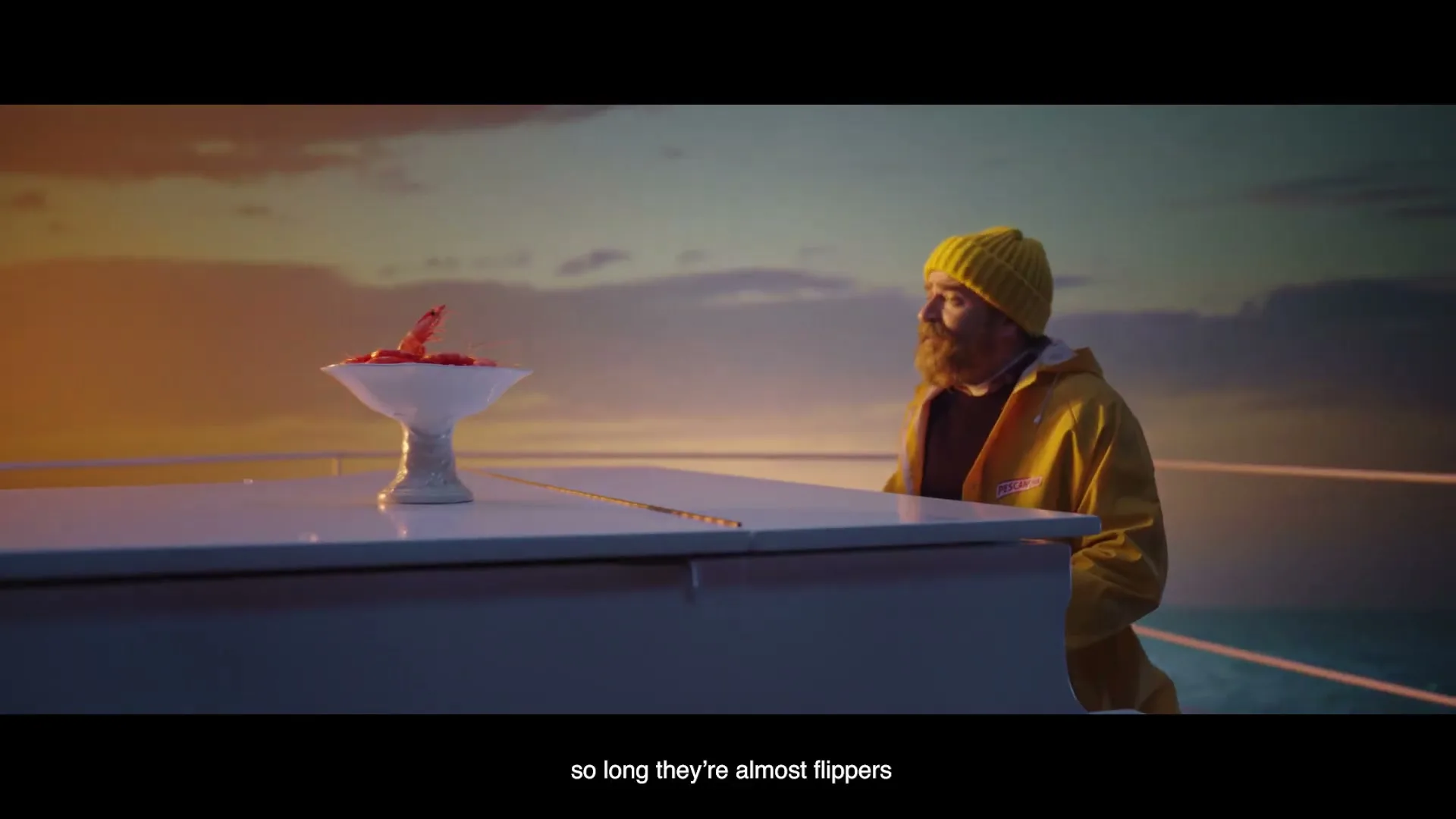 A bearded man in a yellow raincoat and beanie sits at a white piano on a boat, looking at a bowl of shrimp at sunset.