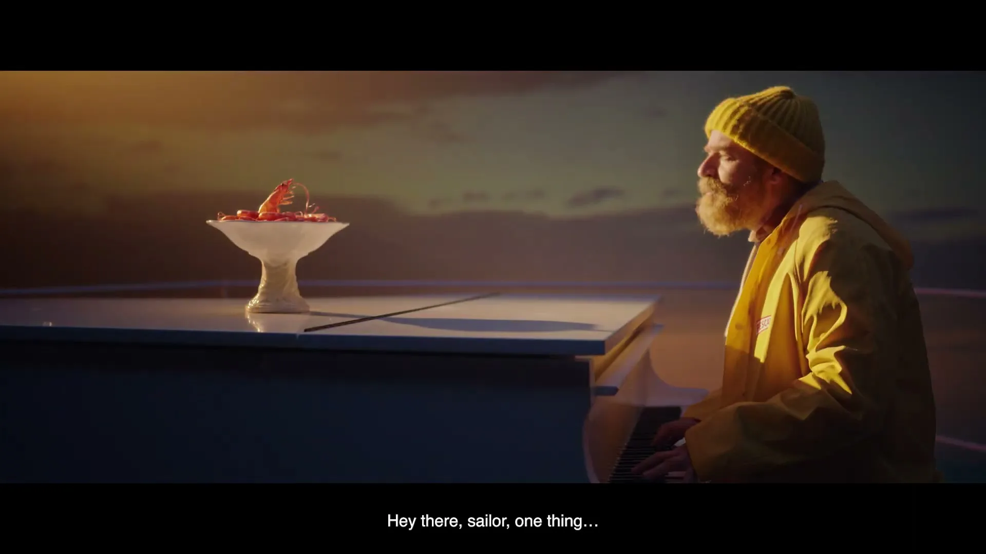 A bearded man in a yellow beanie and raincoat plays a white piano with a bowl of shrimp on it, against a sunset sky.