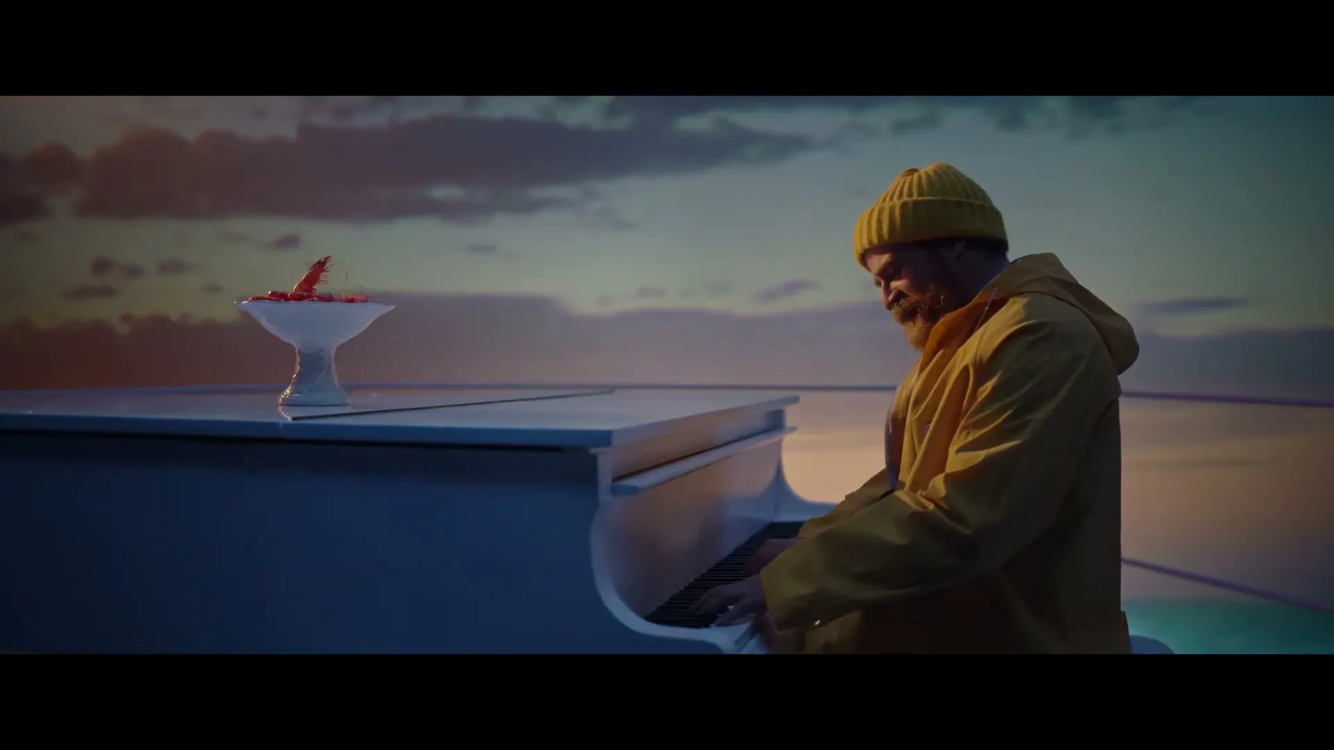 A bearded man in a yellow raincoat and beanie plays a white piano on a boat deck at sunset, with a bowl of red shrimp on the piano.
