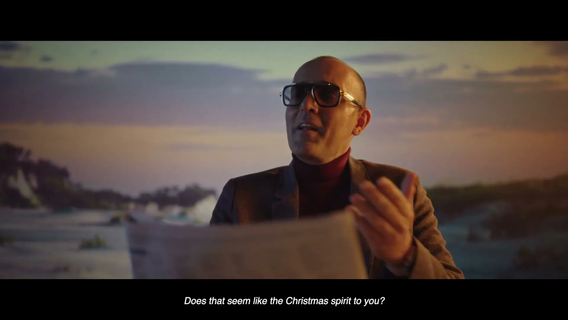 A man in sunglasses and a suit sits at a table, holding a newspaper, with a beach and sunset in the background.
