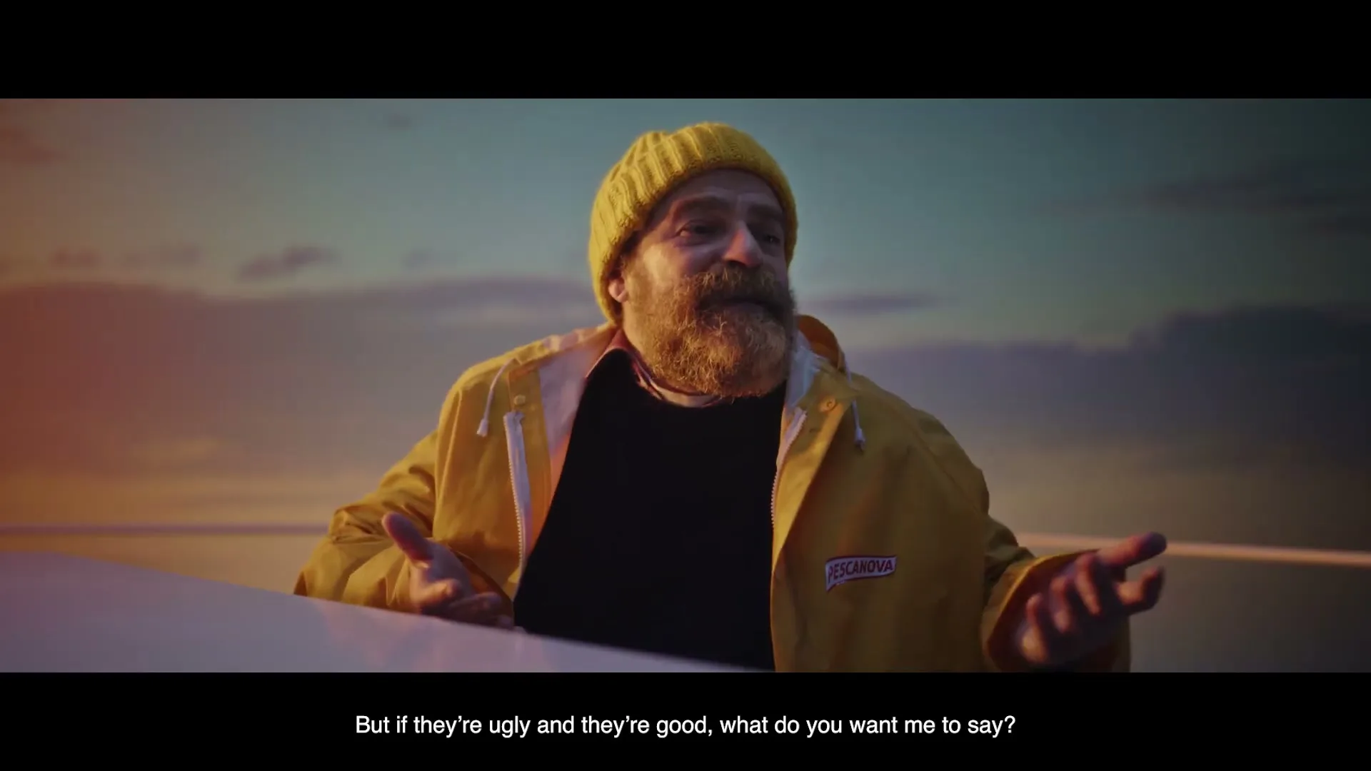 A bearded man in a yellow beanie and raincoat shrugs his shoulders, talking to someone off-camera on a boat at sunset.