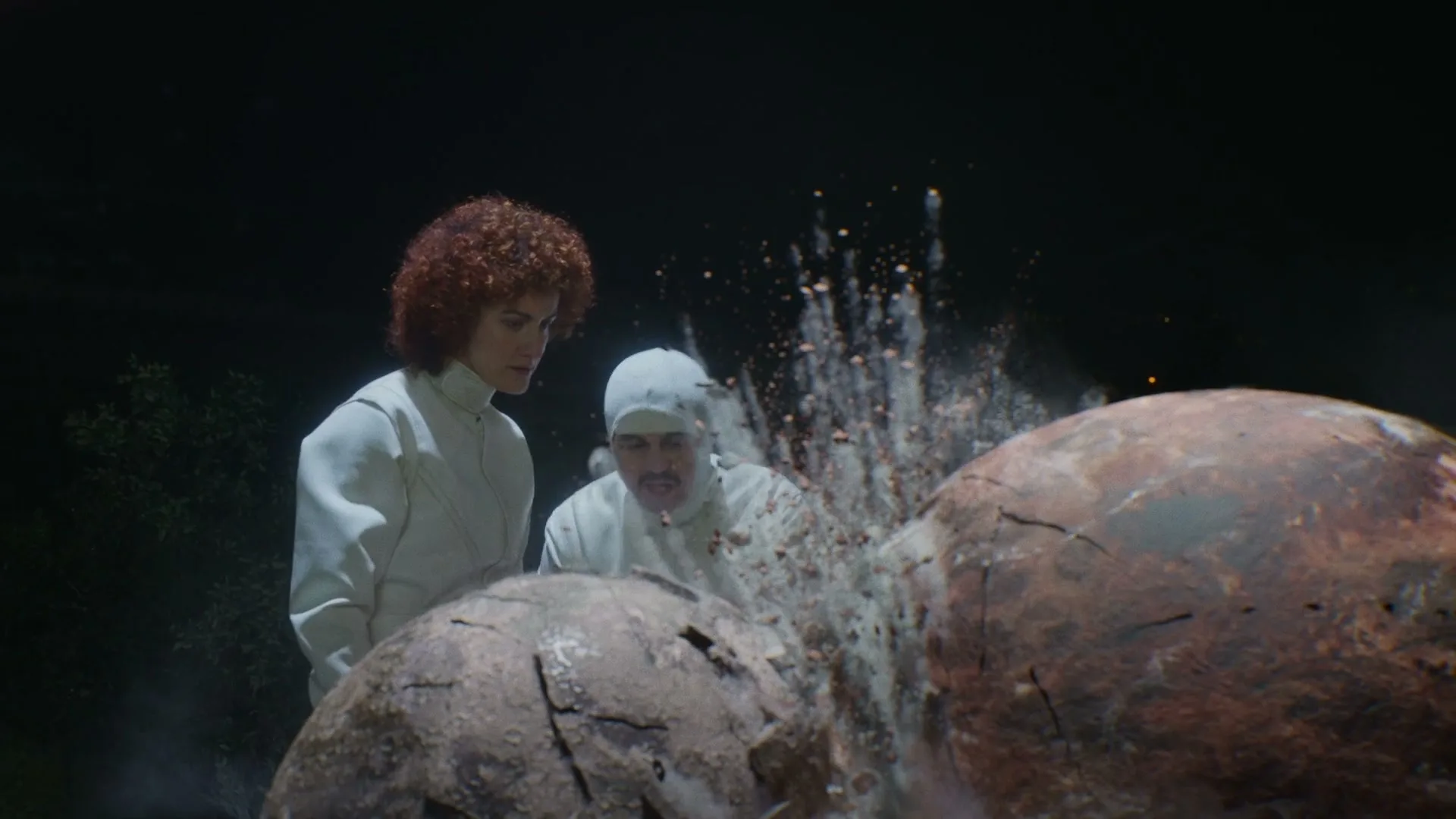 A woman with curly red hair and a man in a white balaclava watch as a large, cracked sphere breaks apart, scattering debris in a dark, mysterious setting.