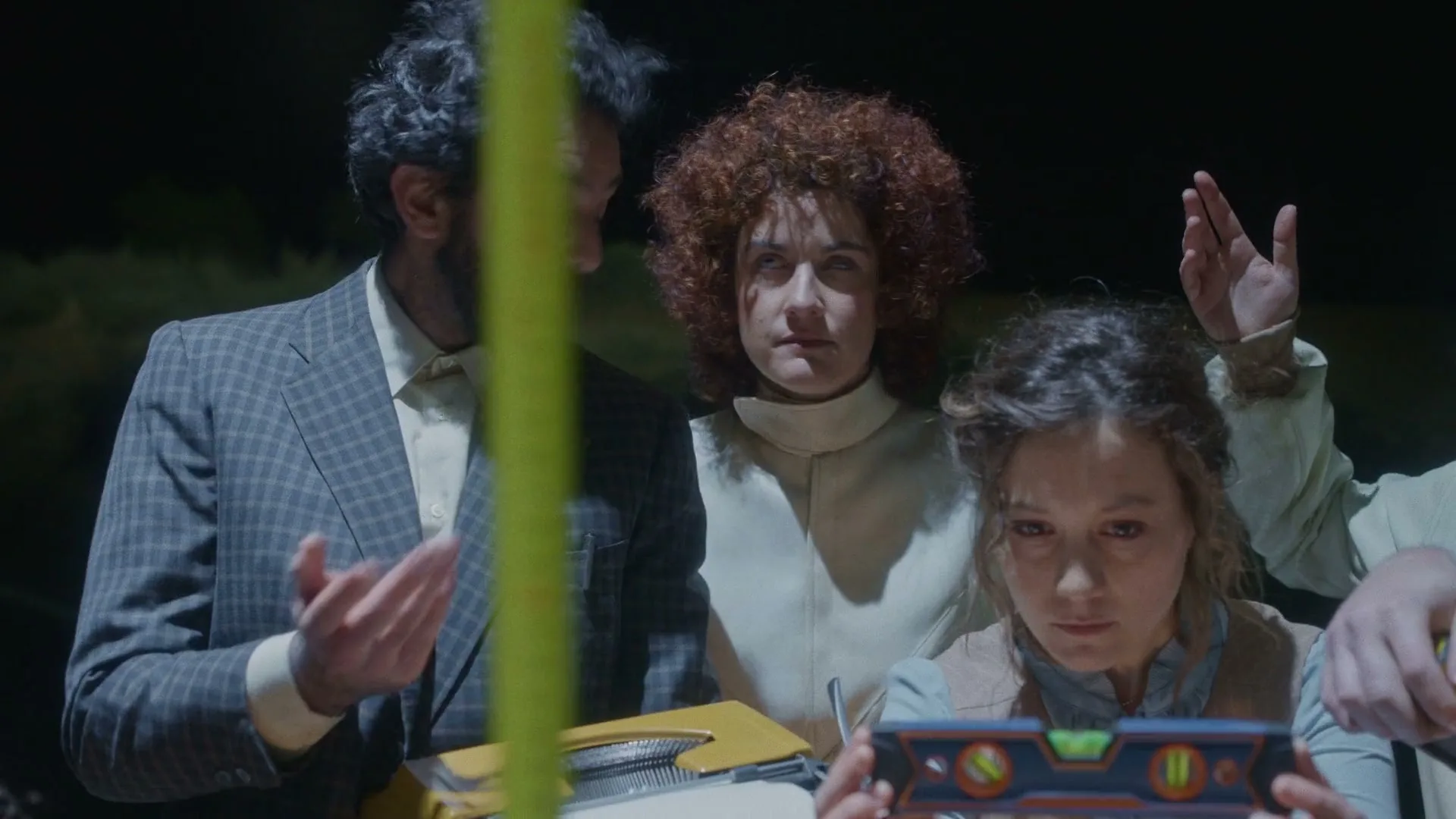 Three people, two women and one man, dressed in period clothing, are looking intently at a device, possibly a level, in a dark, indeterminate setting.