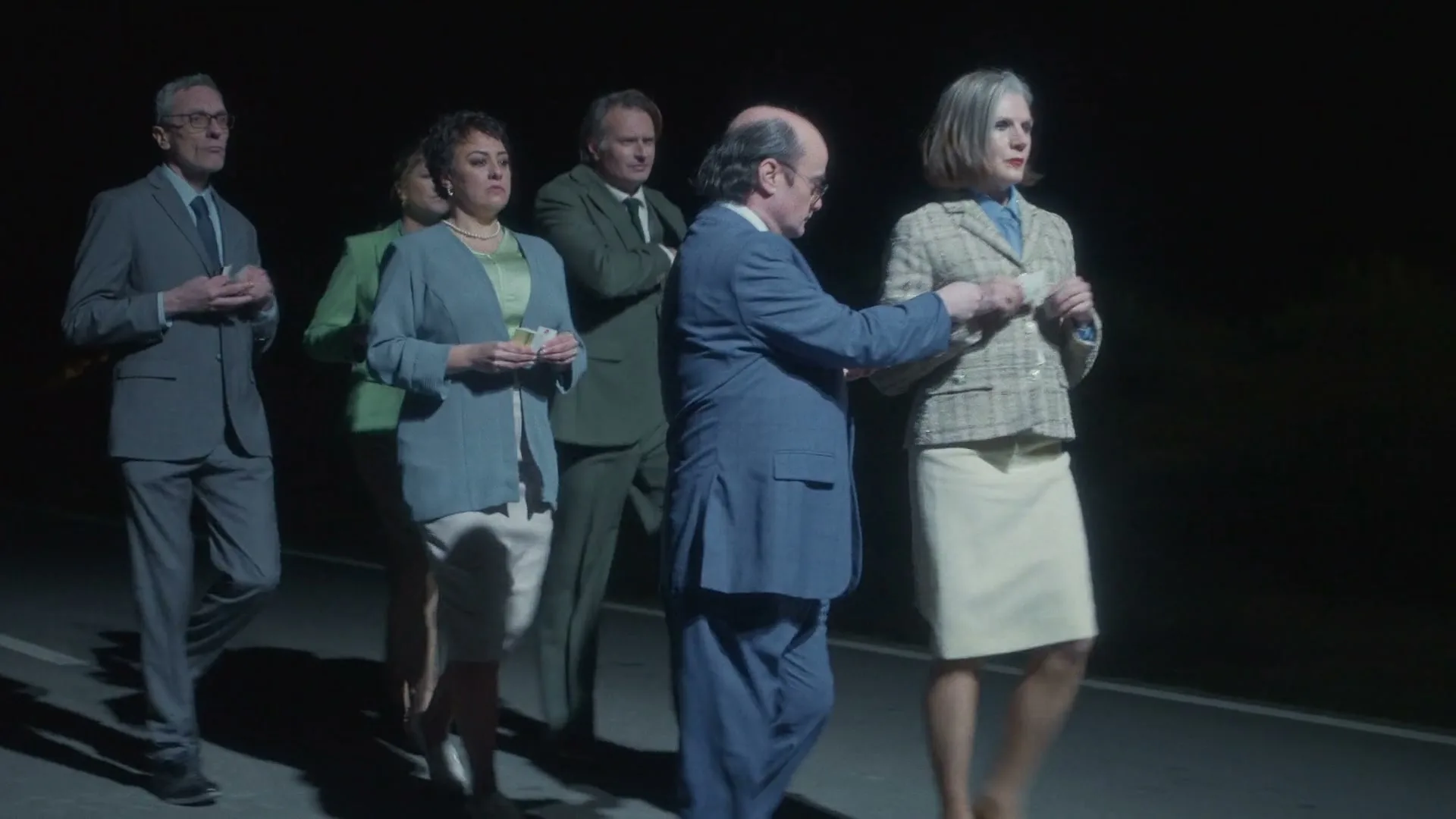 A group of six well-dressed adults, three men and three women, walk together on a dark road at night, looking at small papers in their hands.