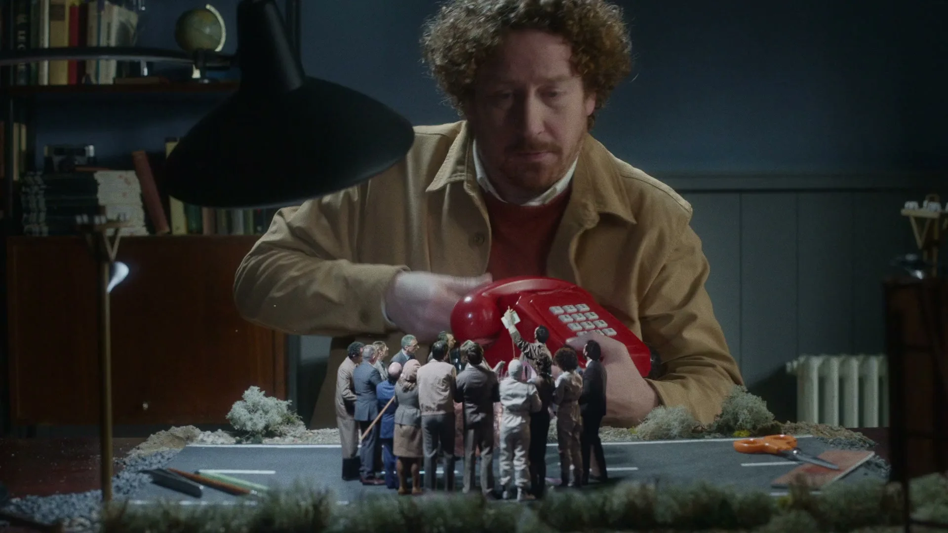 A man with curly hair looks down at a miniature diorama of people gathered around a red telephone on a desk.