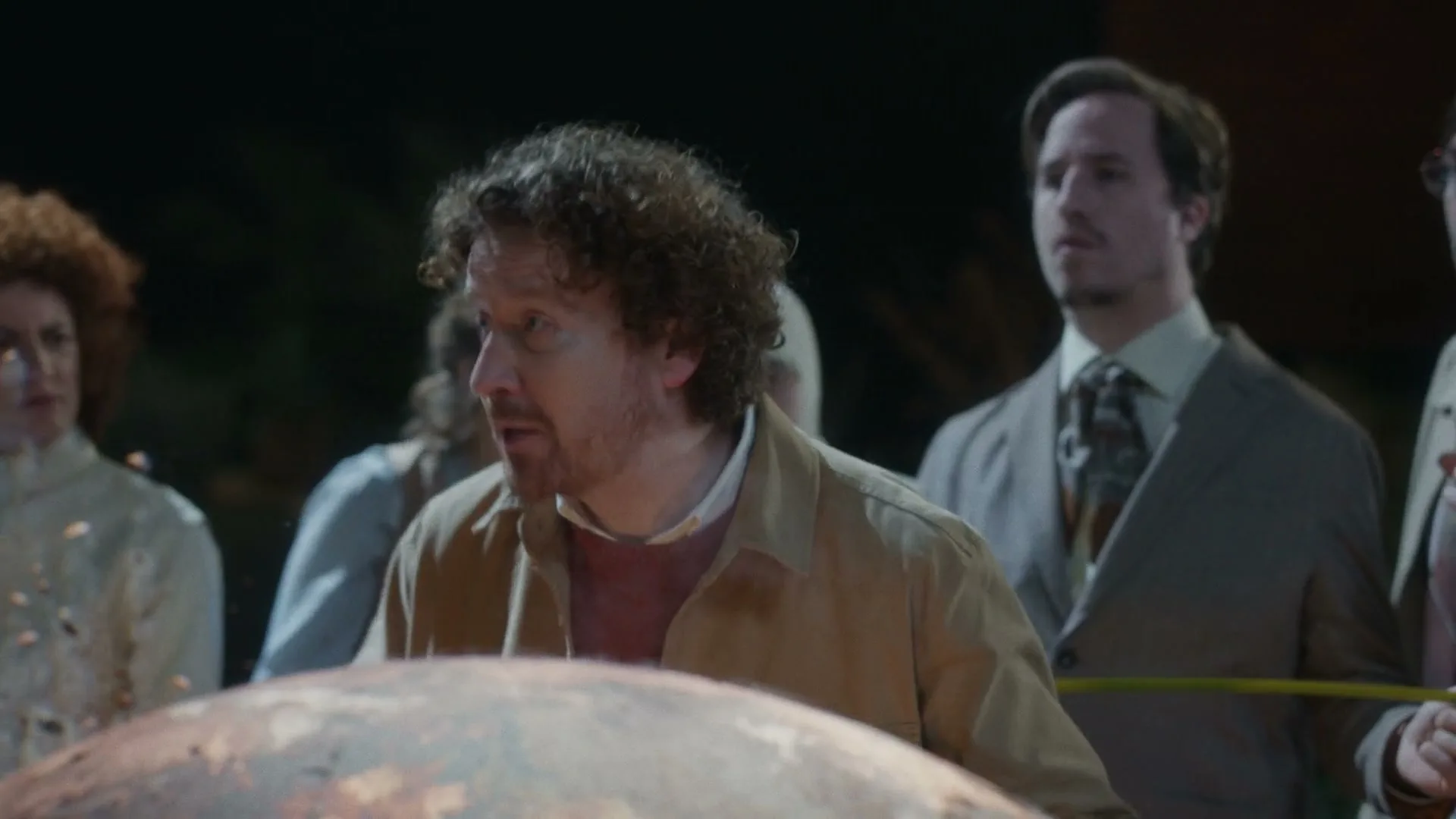 A man with curly hair and a beard looks concerned while gesturing with his hands, surrounded by other people in a dimly lit, indeterminate setting.
