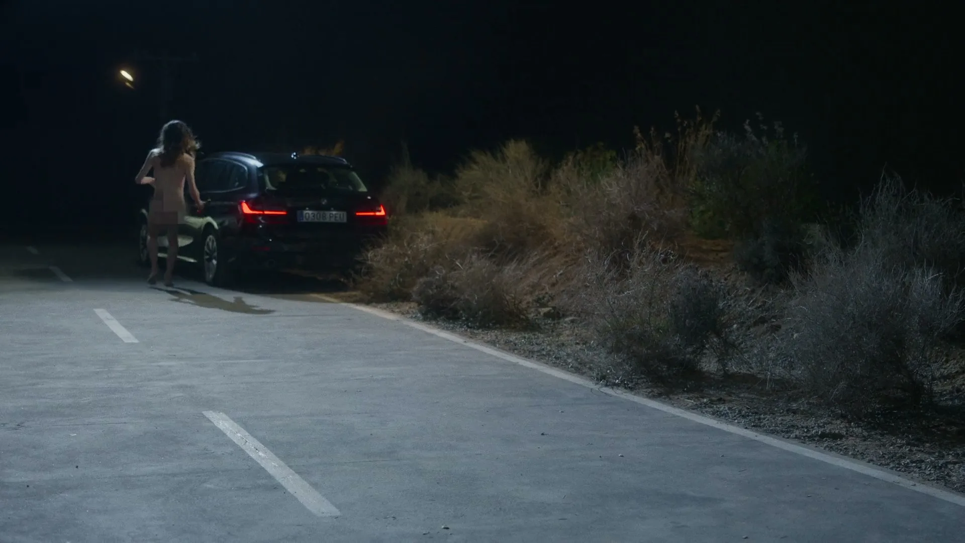 A naked woman with long hair runs towards a black car parked on the side of a dark, deserted road at night.